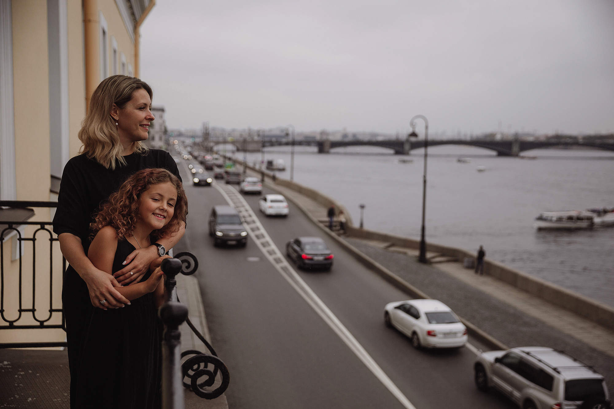 Мама обнимает дочку стоя на балконе на фоне города