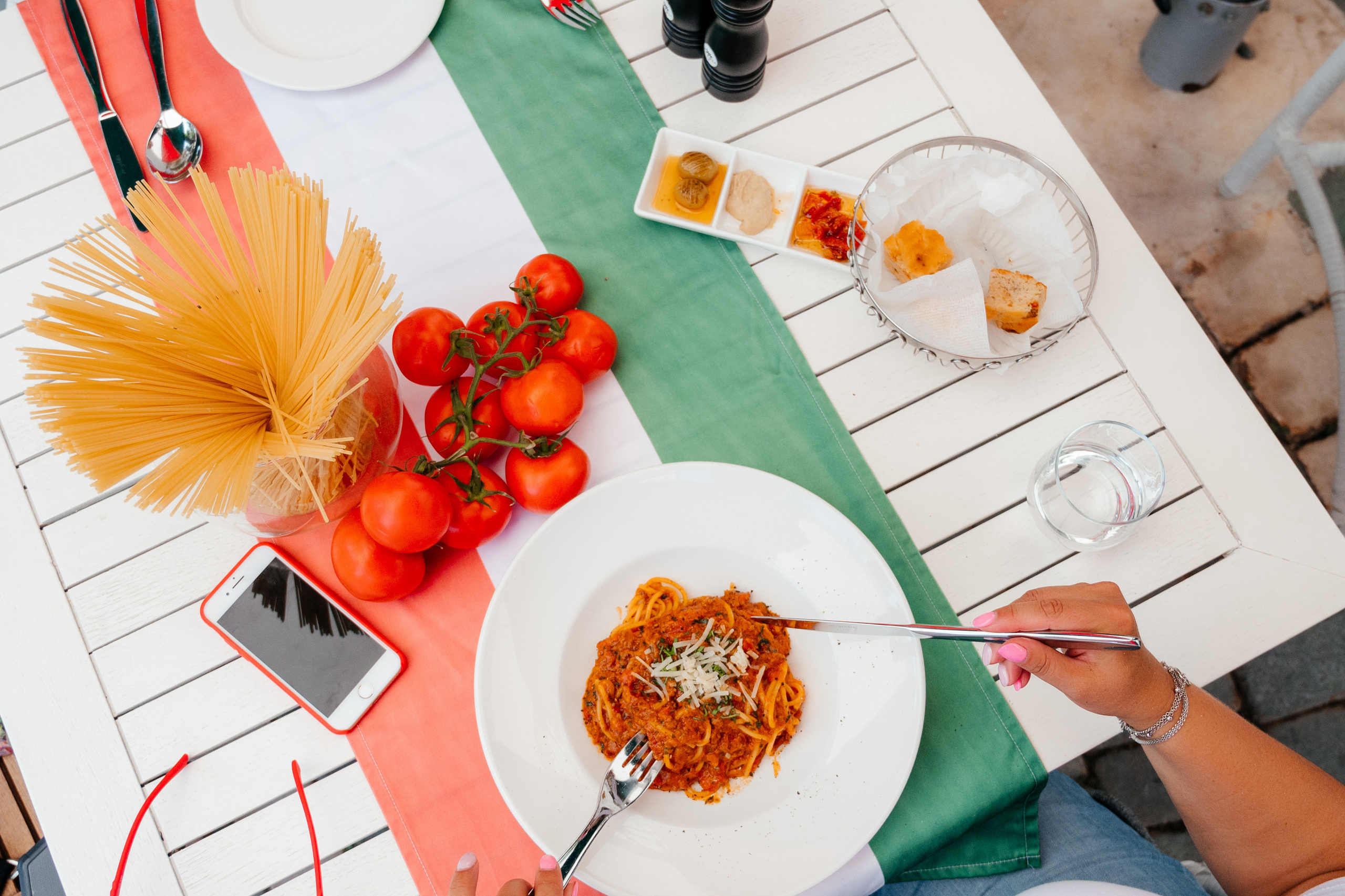 Вид с верху, на столе со скатертью, в виде Итальянского флага, женские руки держат вилку и нож. На вилке Итальянская паста. На столе стоит ваза спастой