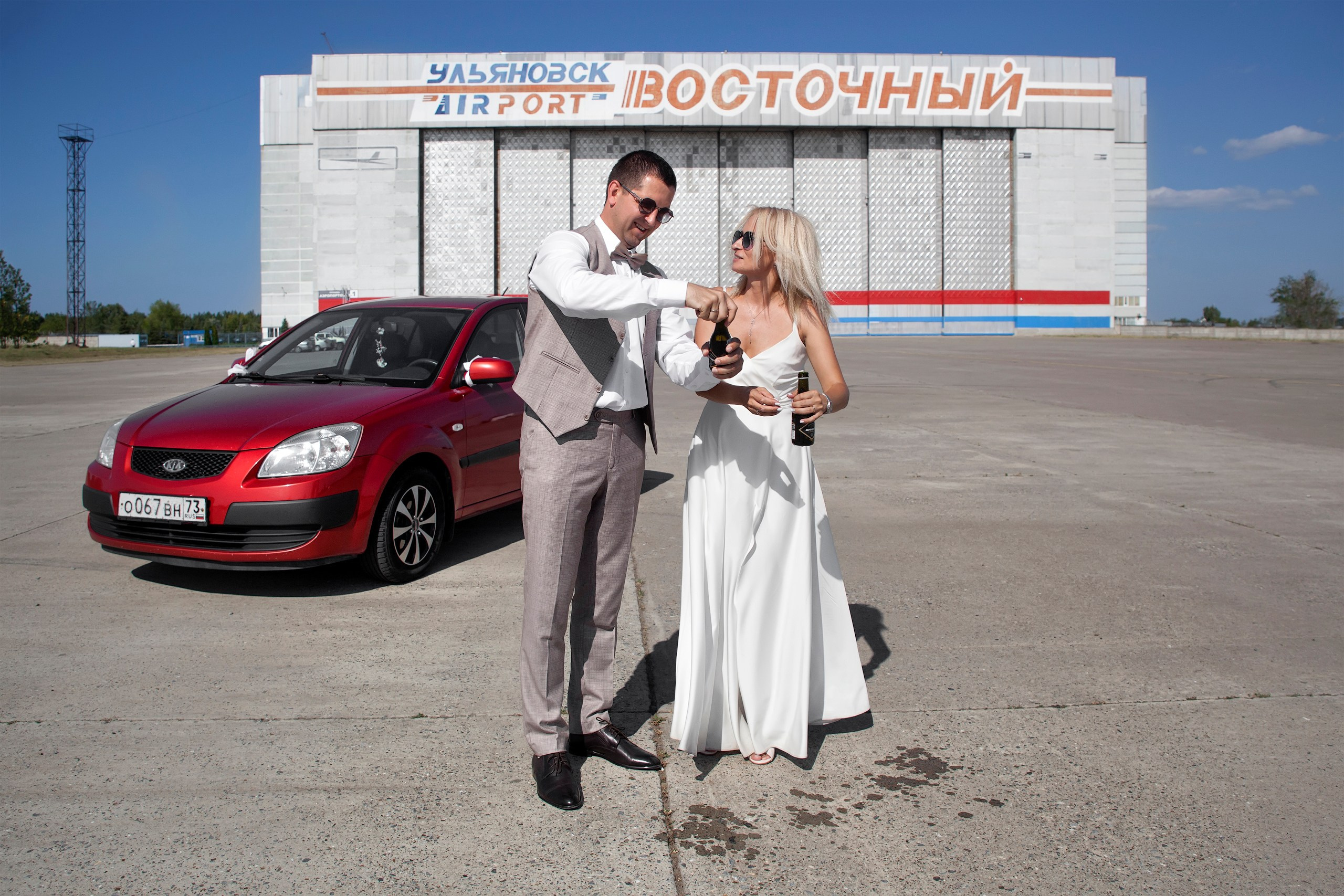 Жених с невестой открывают шампанское в малых бутылочках на фоне аэропорта