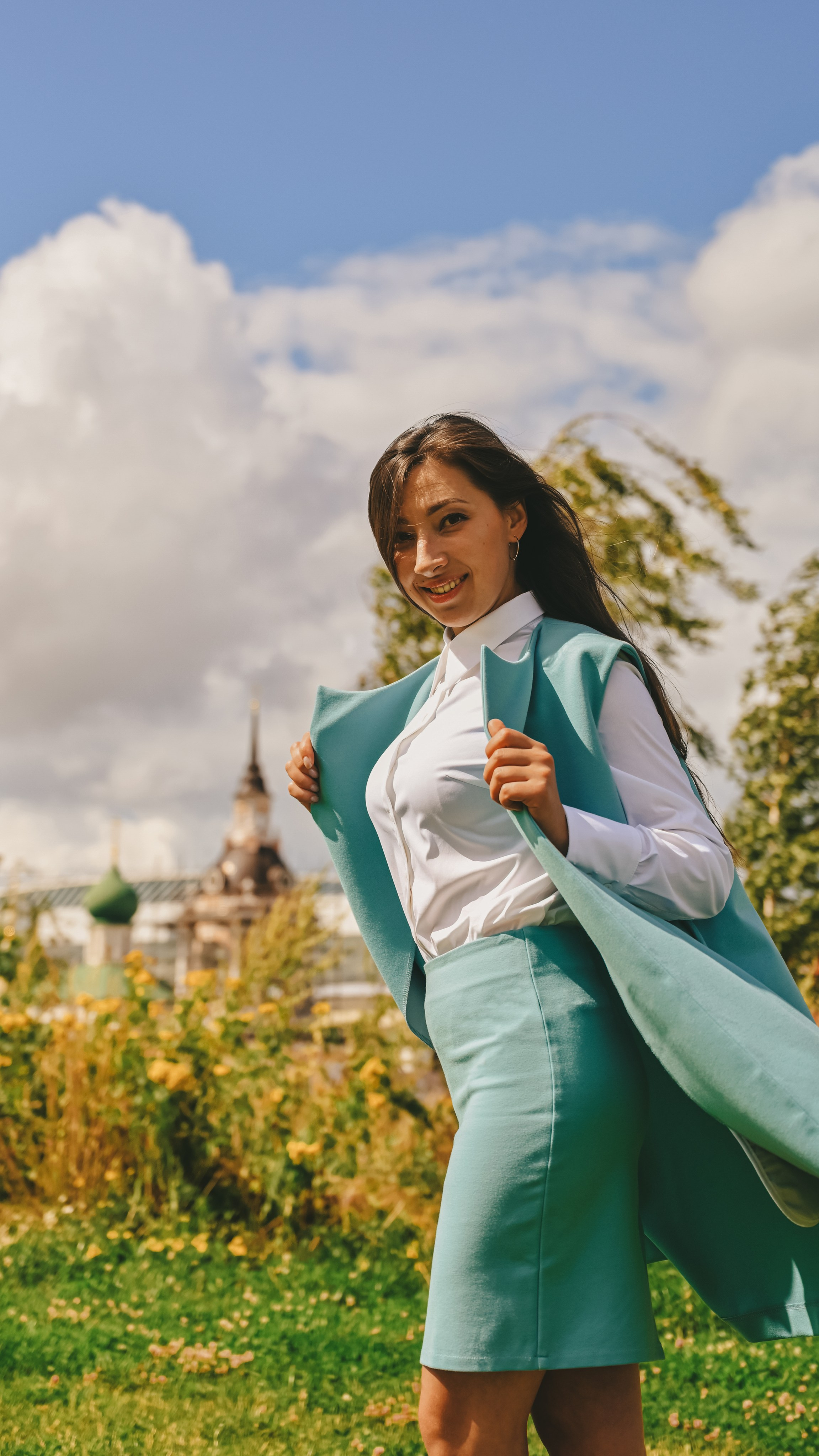Молодая женщина в светло-зеленом костюме стоит на фоне сада и неба. Она одета в белую блузку, зеленый жакет и юбку. Волосы собраны назад, она улыбается и держит жакет за плечи. На заднем плане видны цветущие кусты и часть архитектурного сооружения с башней под голубым небом с облаками.