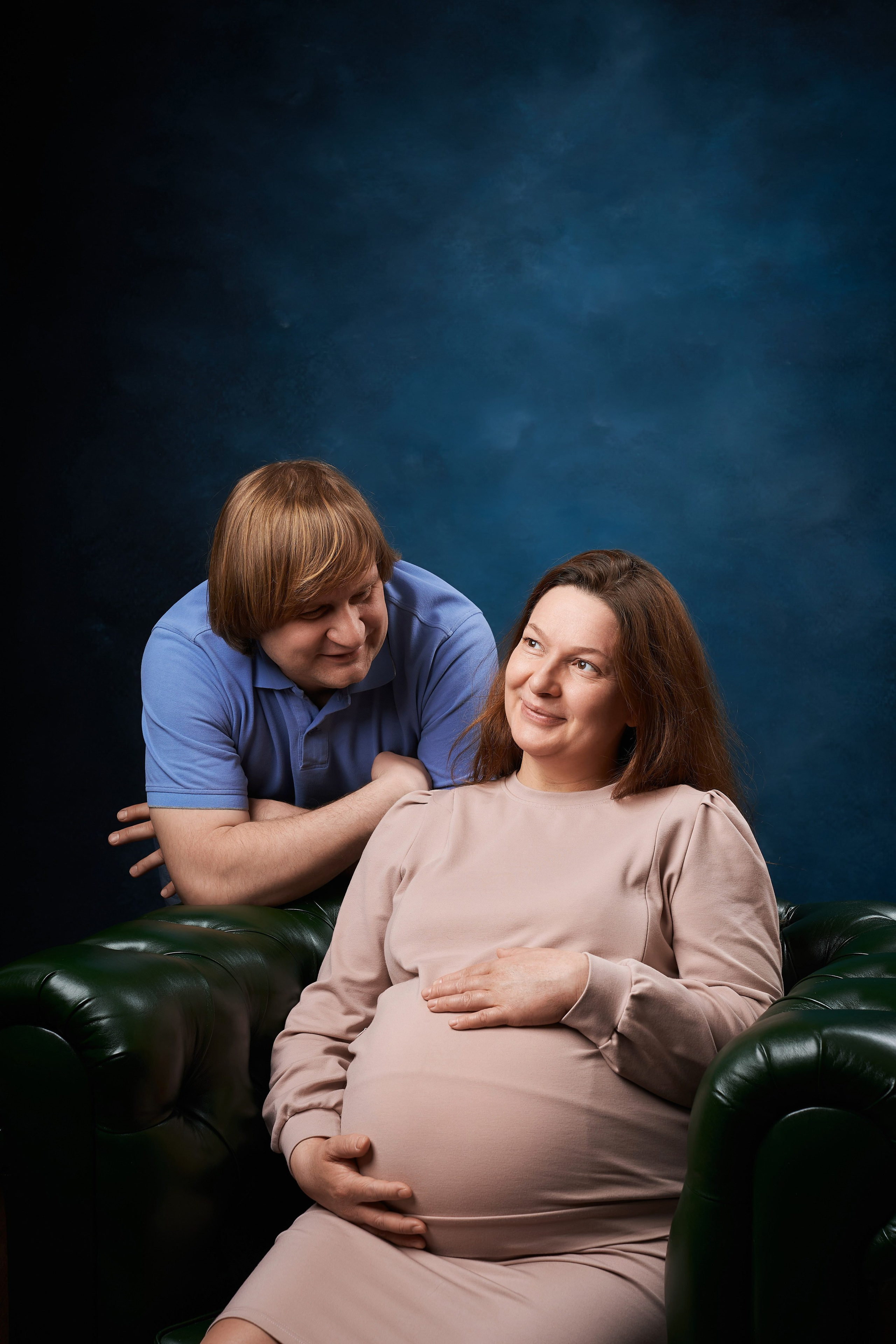 муж смотрит на любимую беременную жену которая сидит на зелёном кожаном кресле