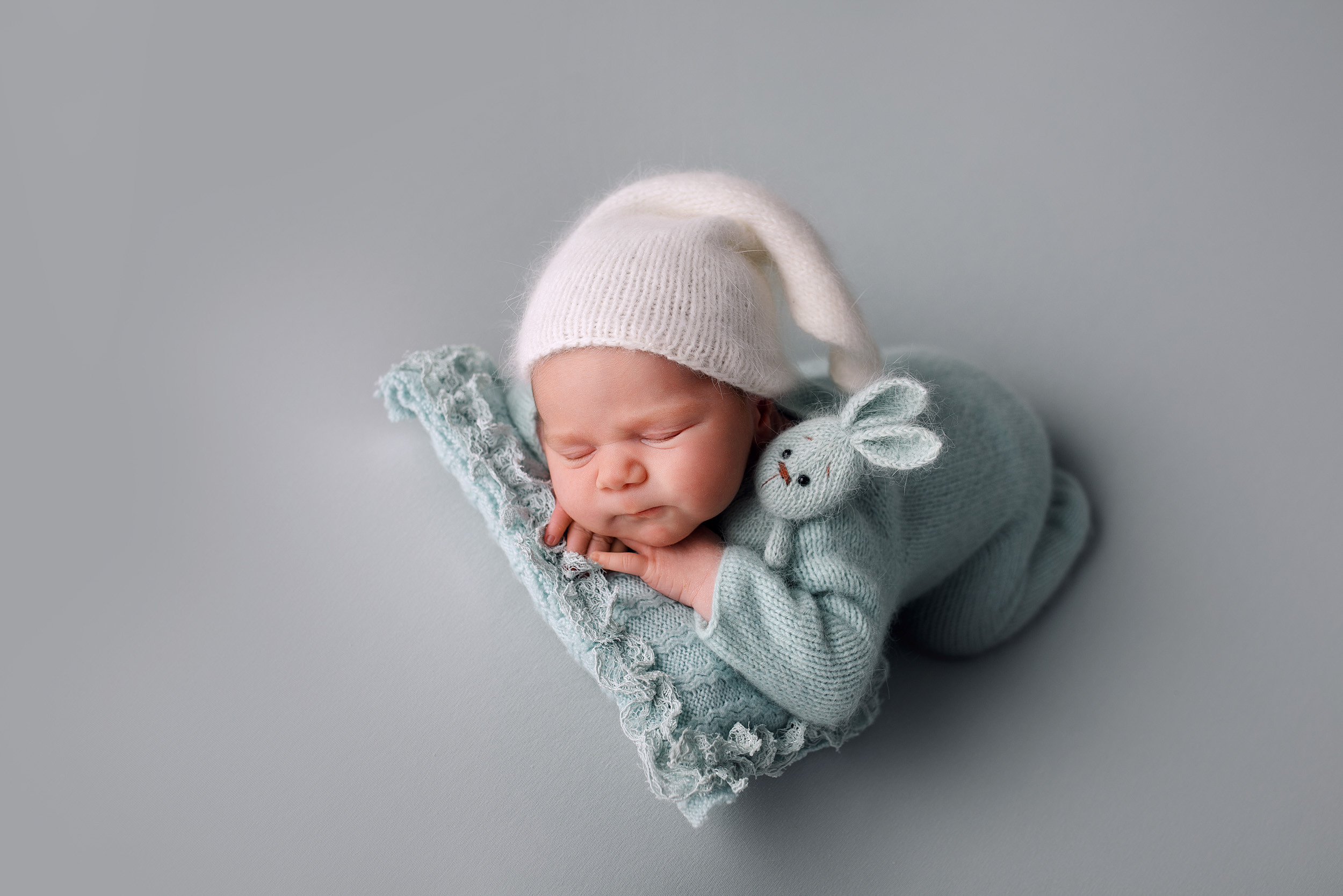 Новорожденный малыш на фотосессии в колпачке с игрушкой позировка ручки под подбородком на фоне
