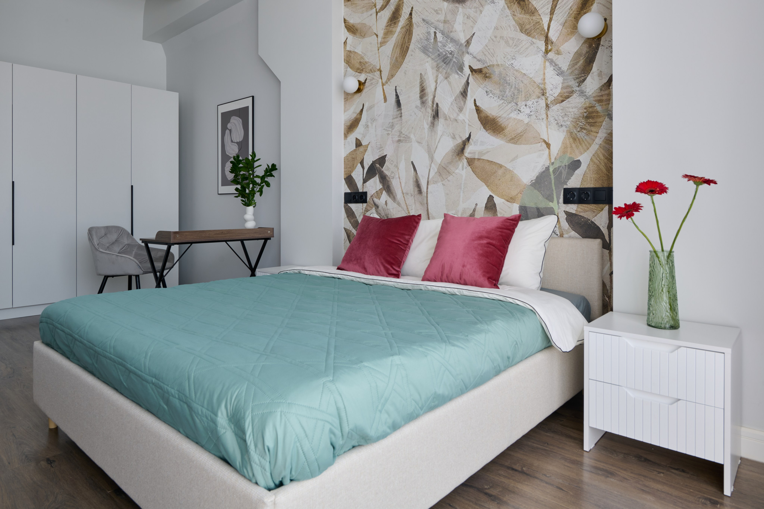 Современная спальня с кроватью, украшенной тюркоазным покрывалом и бордовыми подушками. На заднем фоне видна стена с абстрактным декоративным панно. Справа стоит небольшой белый комод с вазой и красными цветами.