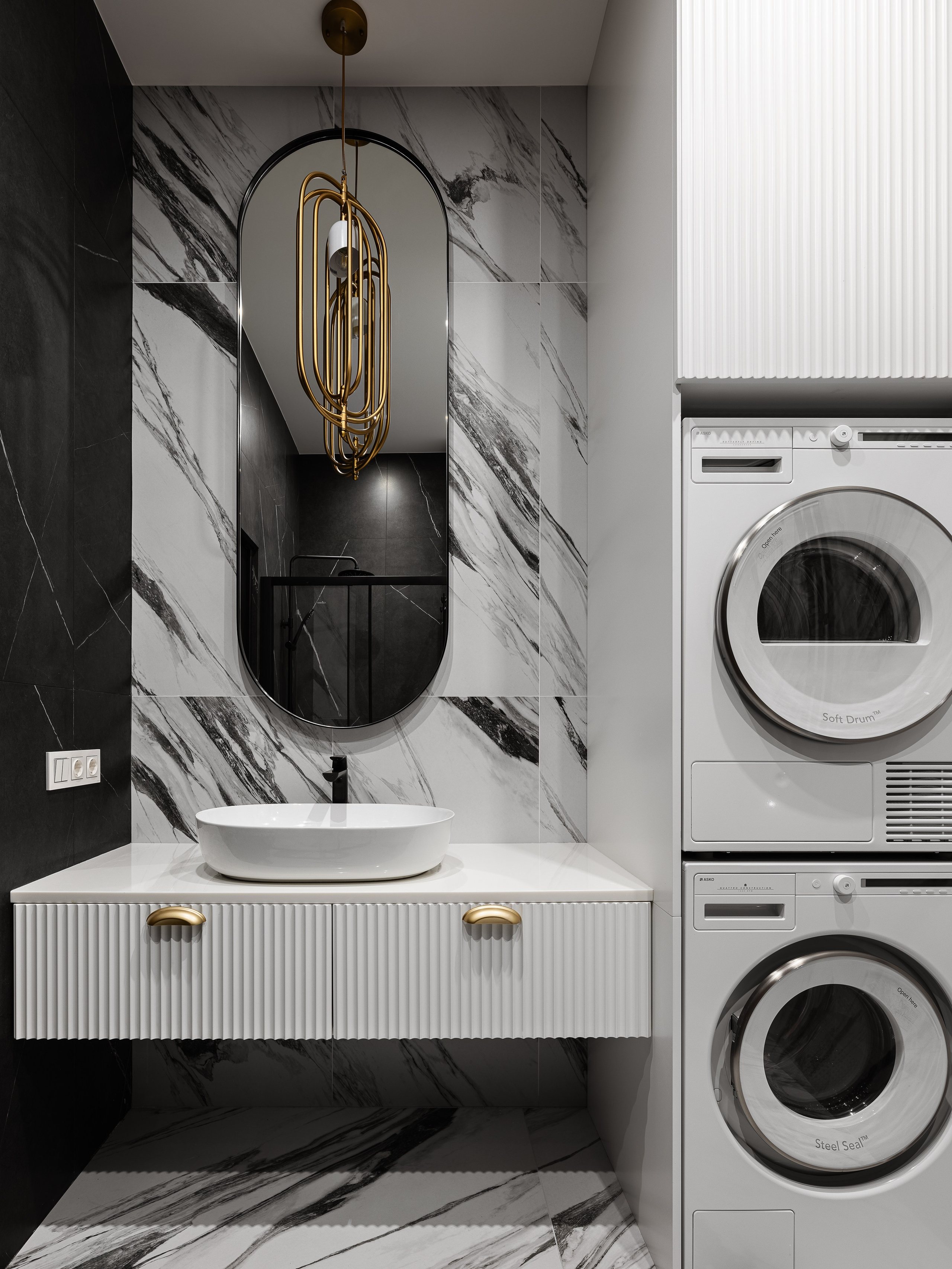 Интерьер ванной комнаты с прачечной зоной: стиральная и сушильная машины в колонне, белая тумба с рифлёными фасадами и золотыми ручками, мраморные плитки и дизайнерское зеркало, автор Василисса Бачева, стилист Наталья Якушева, фото Сергей Денисенко.