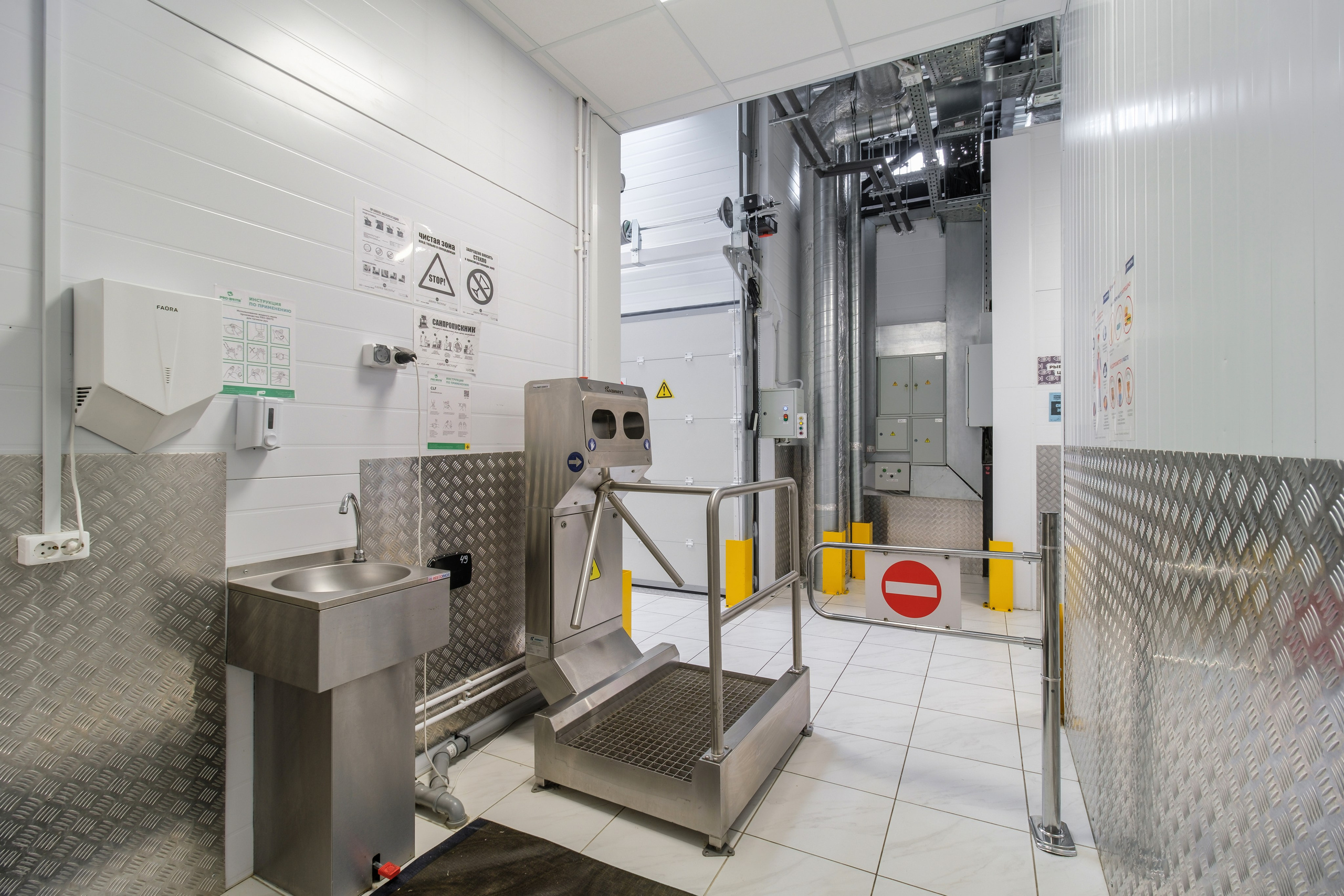 Проходная в промышленном здании. В центре кадра находится турникет со встроенной мойкой рук. Пол покрыт плиткой, а стены и потолок окрашены в светлые цвета.