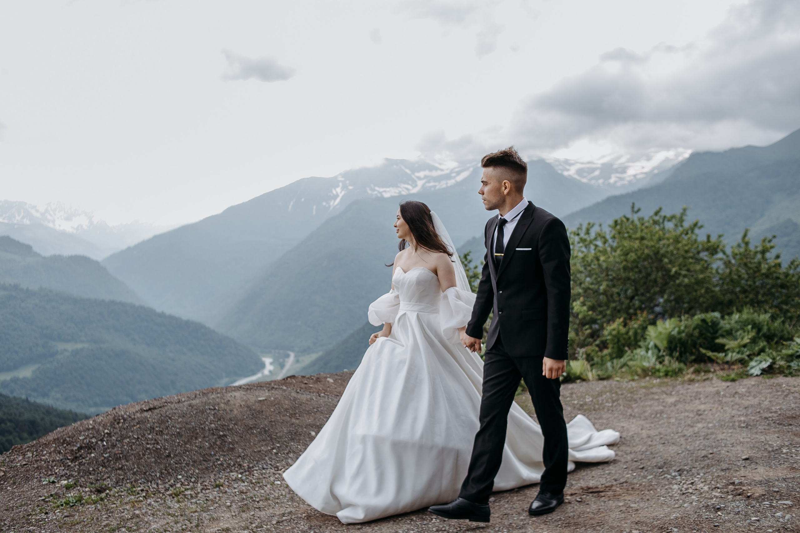 Невеста в белом свадебном платье, стоящая на фоне заснеженных вершин Кавказа с цветами в руках