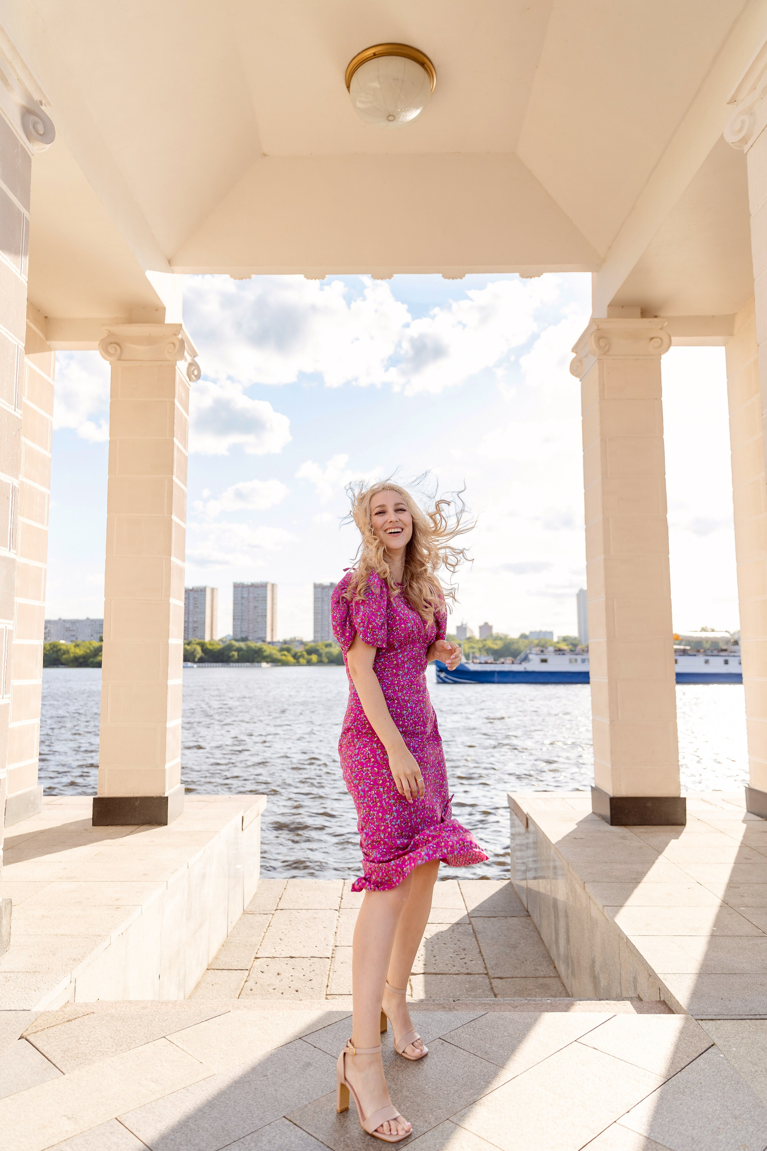 Девушка в розовом платье с развивающимися от ветра волосами стоит у речного вокзала