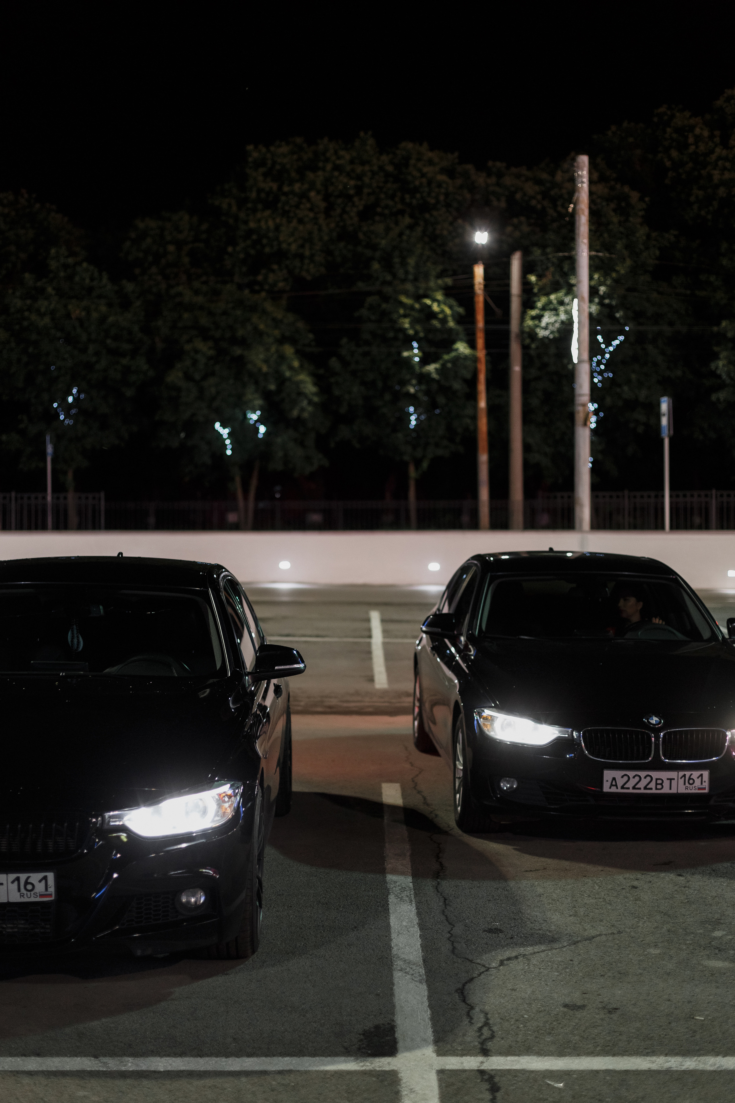 Два чёрных автомобиля BMW на парковке ночью рядом с ТРЦ Мармелад в Таганроге. Фон освещён уличными фонарями и деревьями с подсветкой.