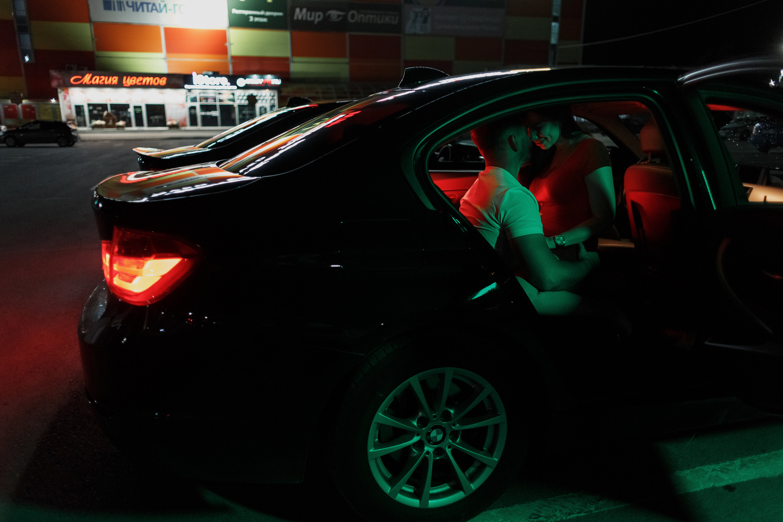 Пара сидит в автомобиле BMW на парковке у ТРЦ Мармелад в Таганроге ночью, освещённая красными и зелёными огнями от светодиодной лампы Yongnuo YN360 III. На заднем плане виден магазин 'Магия цветов'.