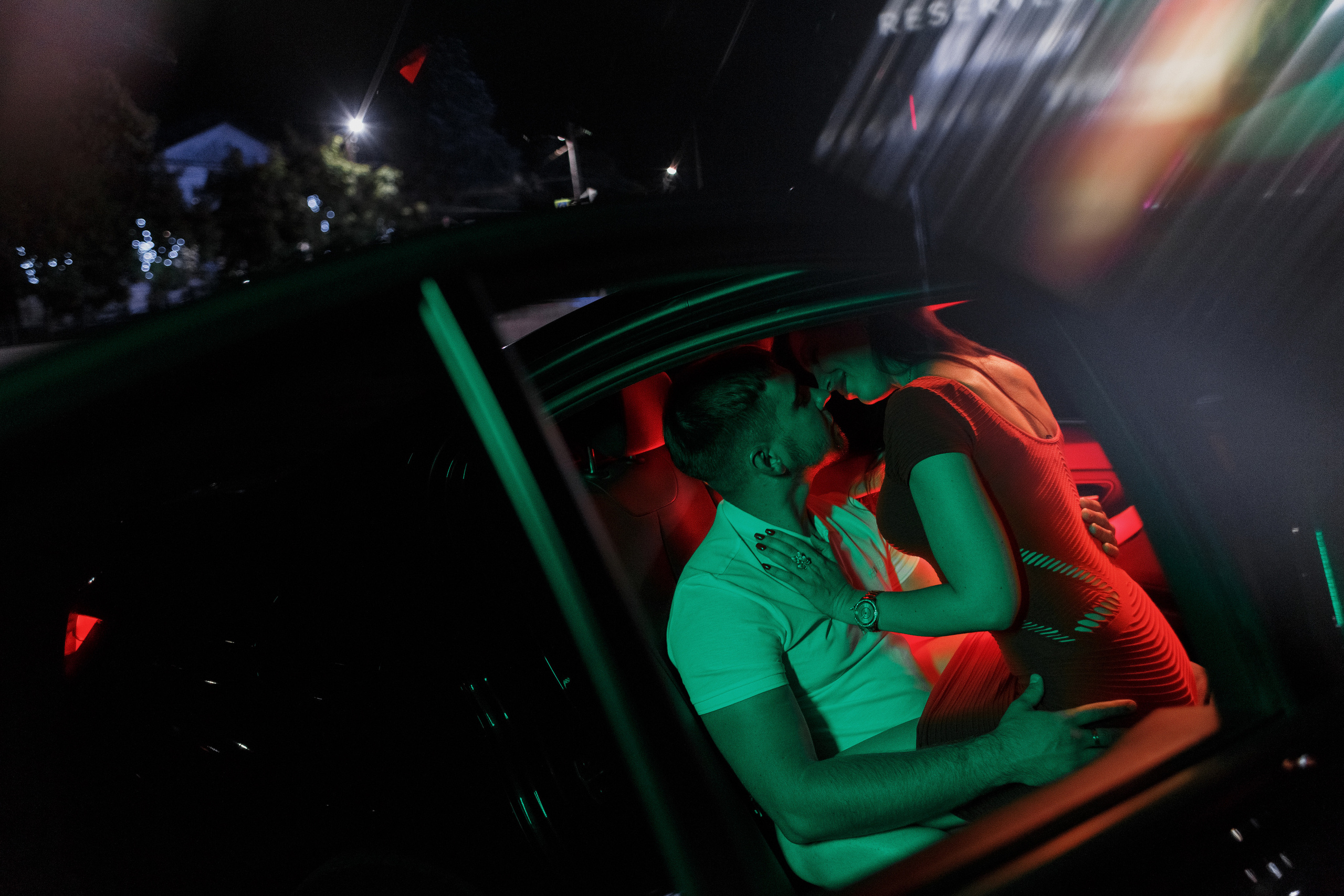 Пара сидит в автомобиле BMW ночью, освещённая светодиодной лампой Yongnuo YN360 III красного и зелёного цвета. Парковка у ТРЦ Мармелад в Таганроге видна на заднем плане.