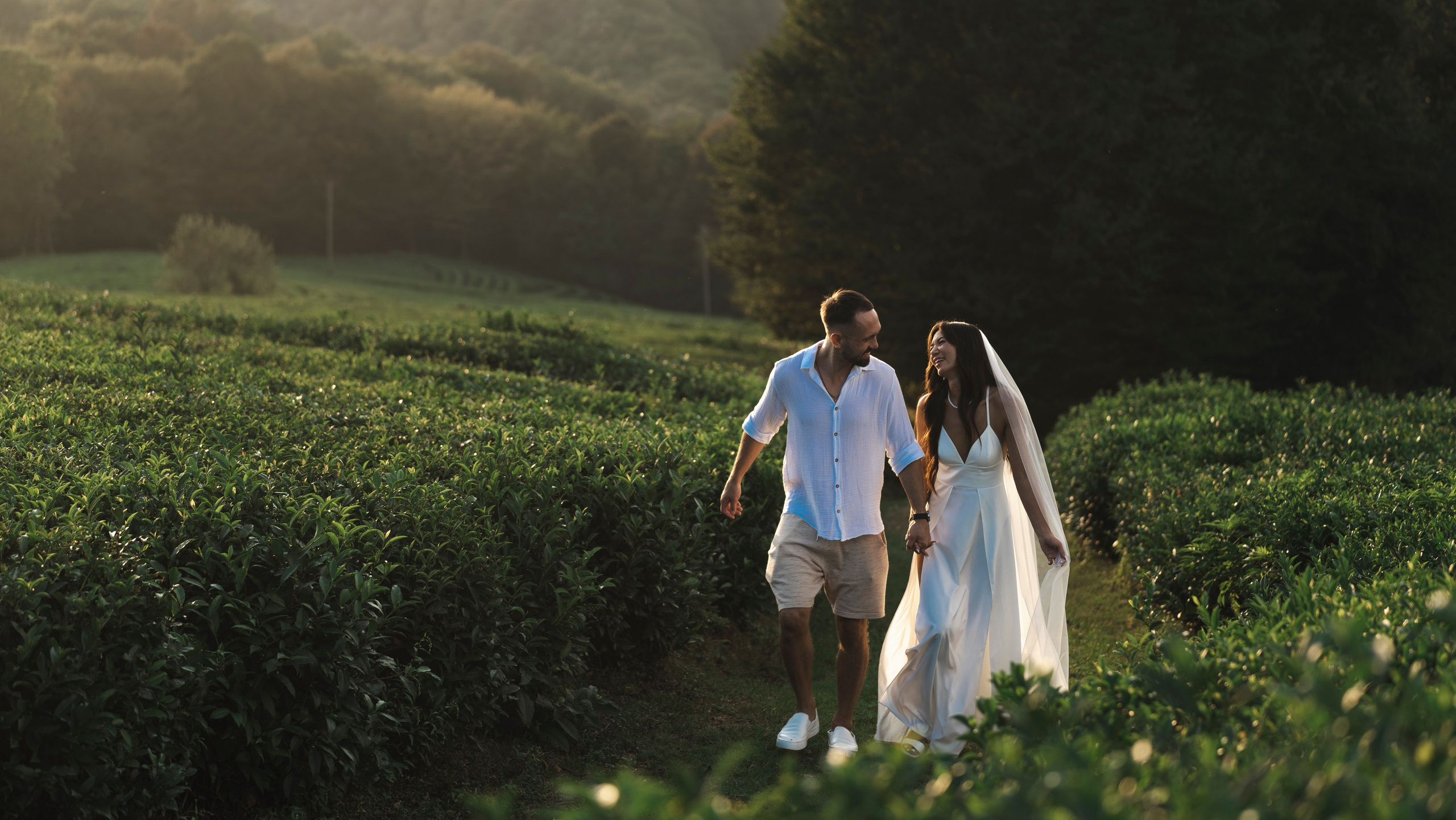 Мужчина и женщина прогуливаются по пышному зеленому полю фермы, держась за руки. На женщине белое платье, а на мужчине белая рубашка и шорты цвета хаки. На заднем плане садится солнце.