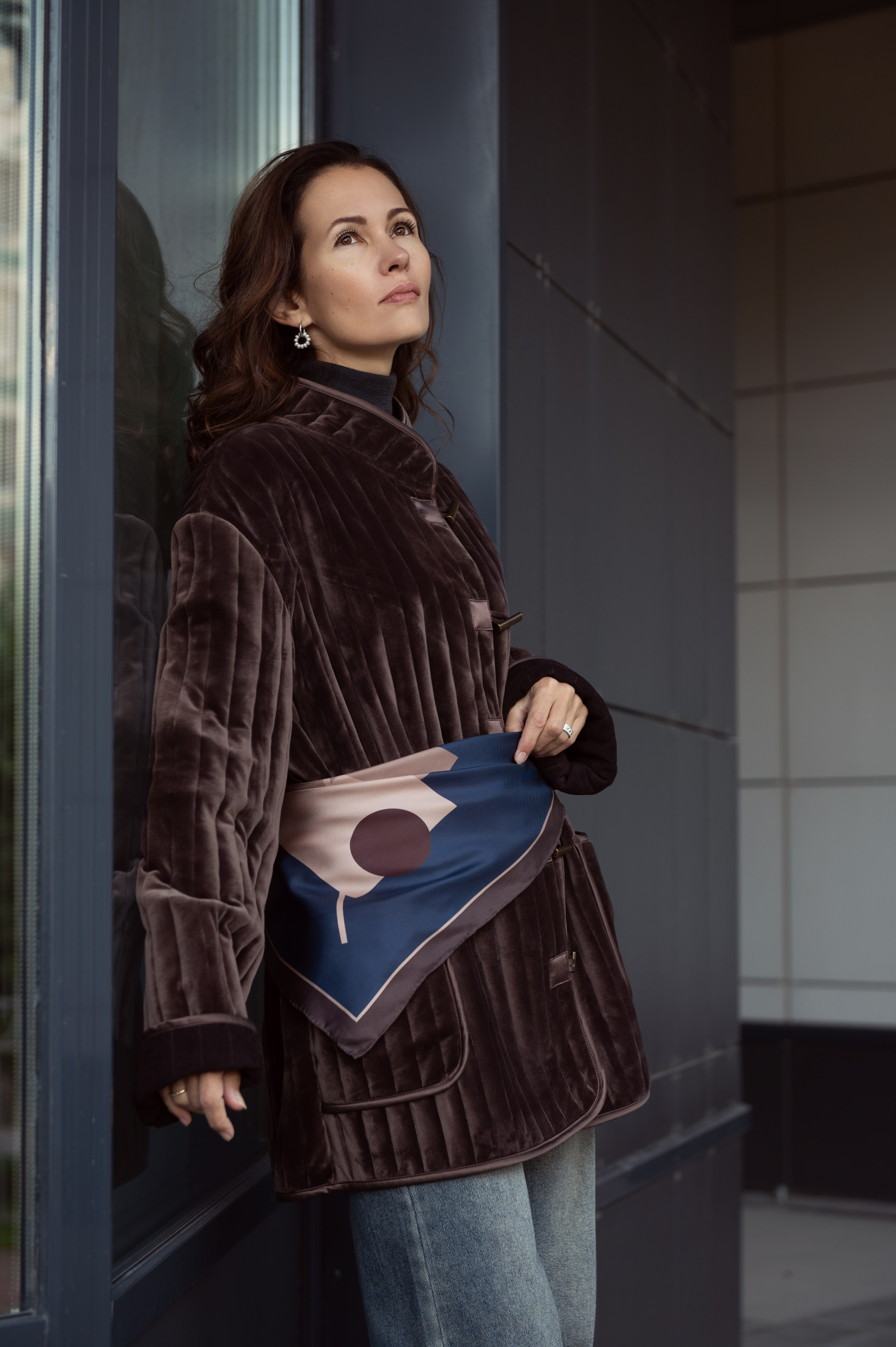 Темноволосая девушка стоит прислонившись к витрине. Съемка для магазина женской одежды и аксессуаров. Естественный свет, мягко отражаясь от стен, окутывает модель и создает равномерно освещенную картинку с красивыми бликами на одежде.