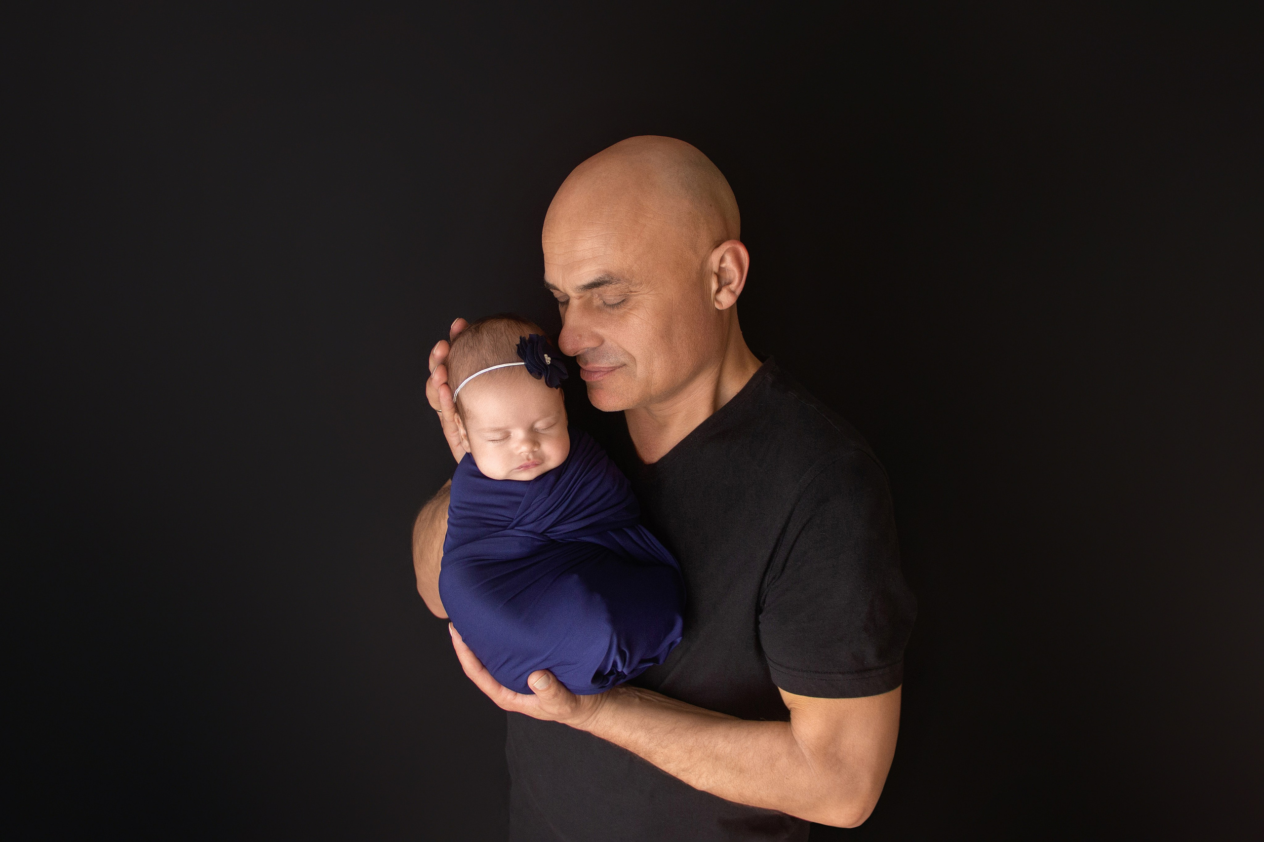 спящая новорожденная девочка с повязочкой на голове лежит в обмотке на сильных руках лысого папы стоящего на черном