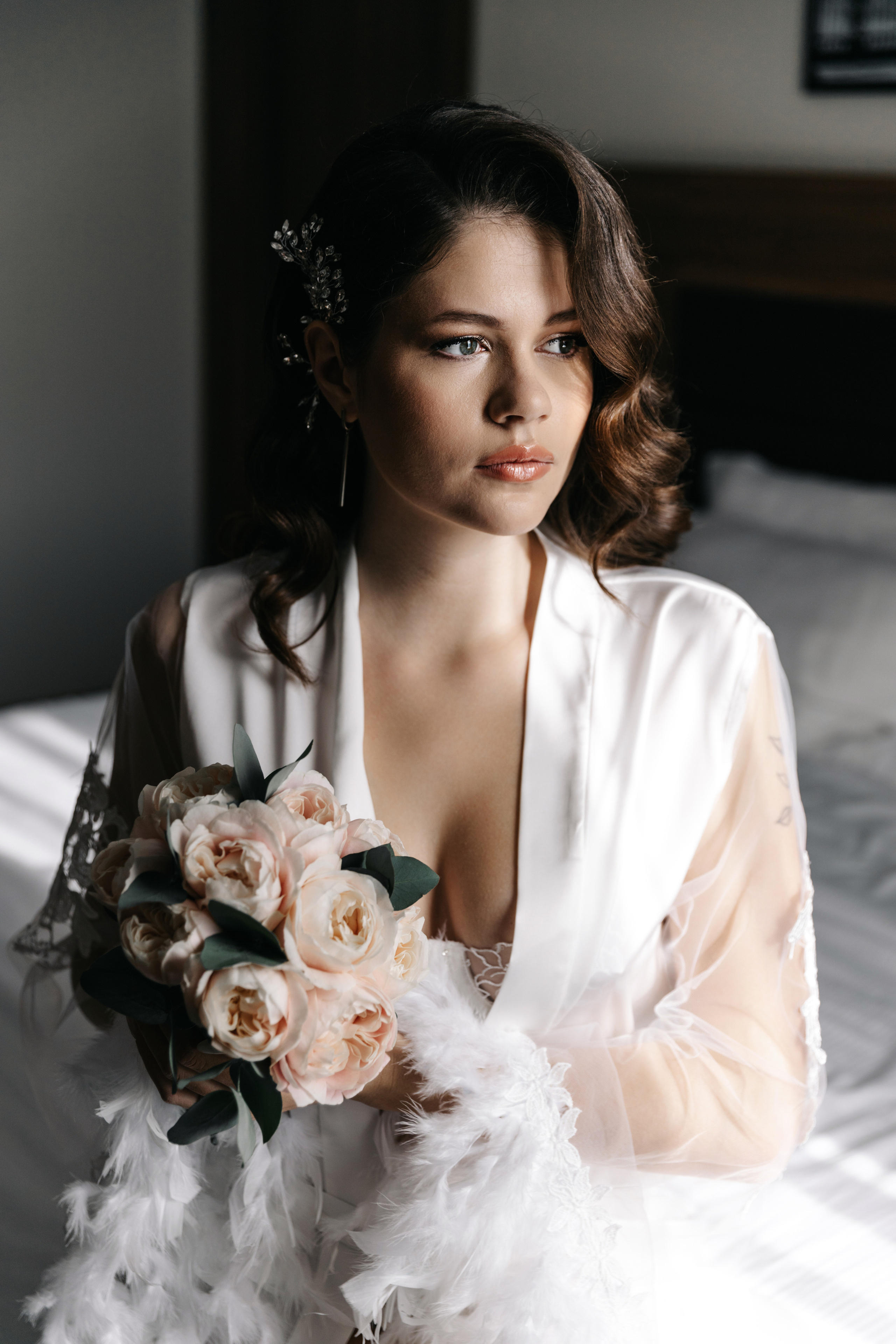 невеста с красивыми локонами смотрит в окно, в руках букет 