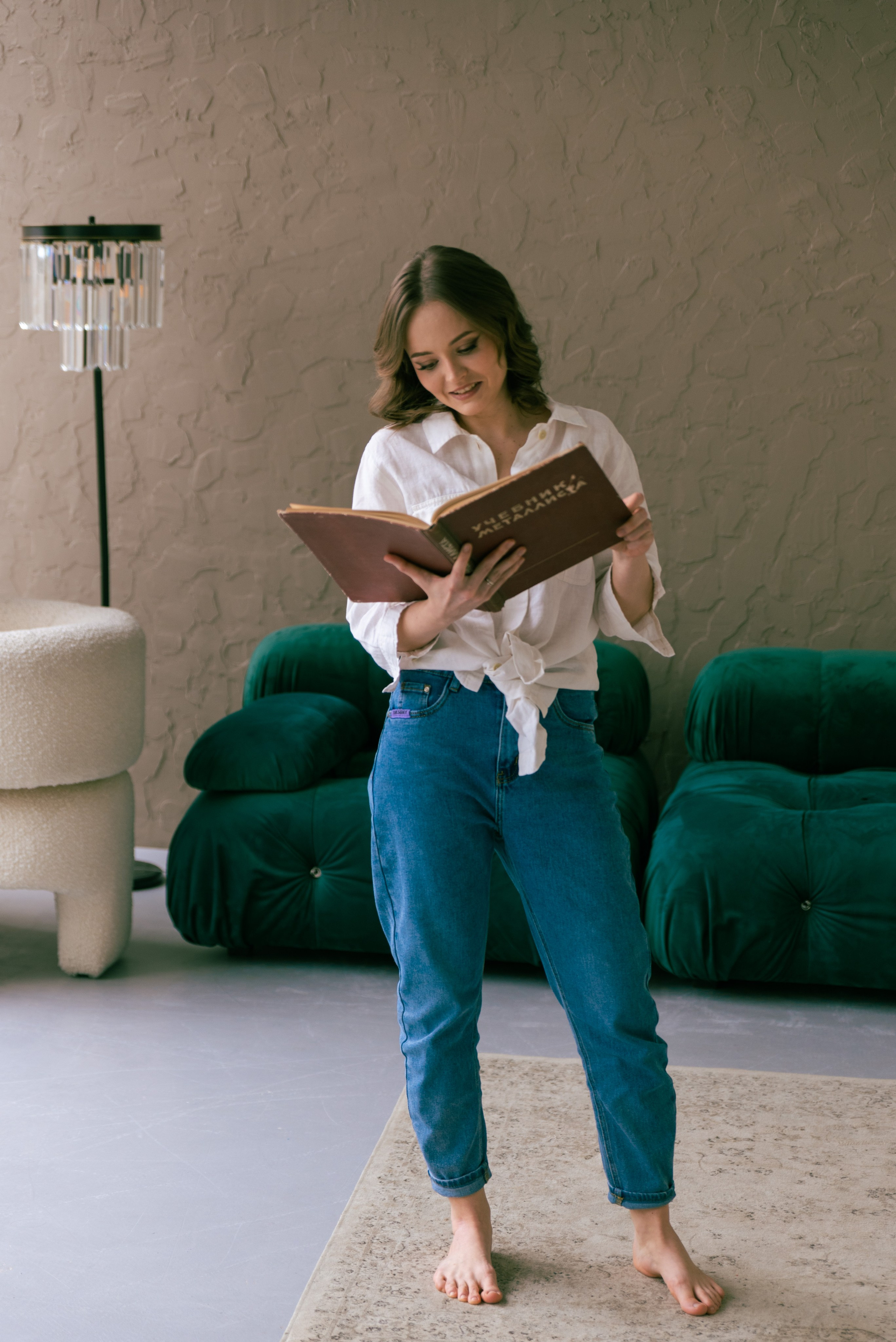 Девушка стоит в комнате, и листает книгу. За ней зелёный диван слева торшер.