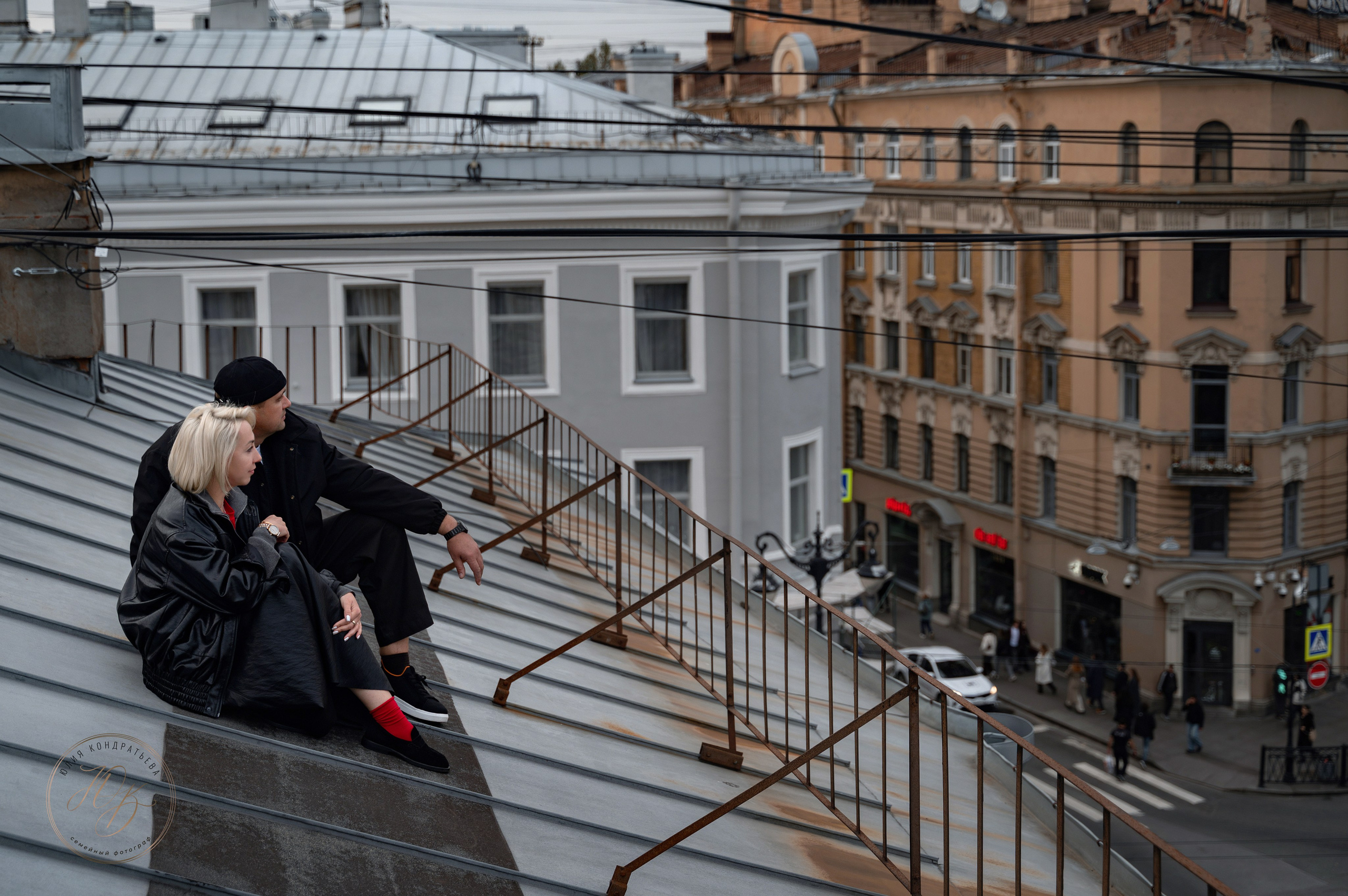 Пара сидит на скате крыши в Петербурге: женщина в чёрной куртке и ярких носках, мужчина в чёрной одежде и шапке. На фоне — исторические здания и городские провода. Атмосферный городской кадр.