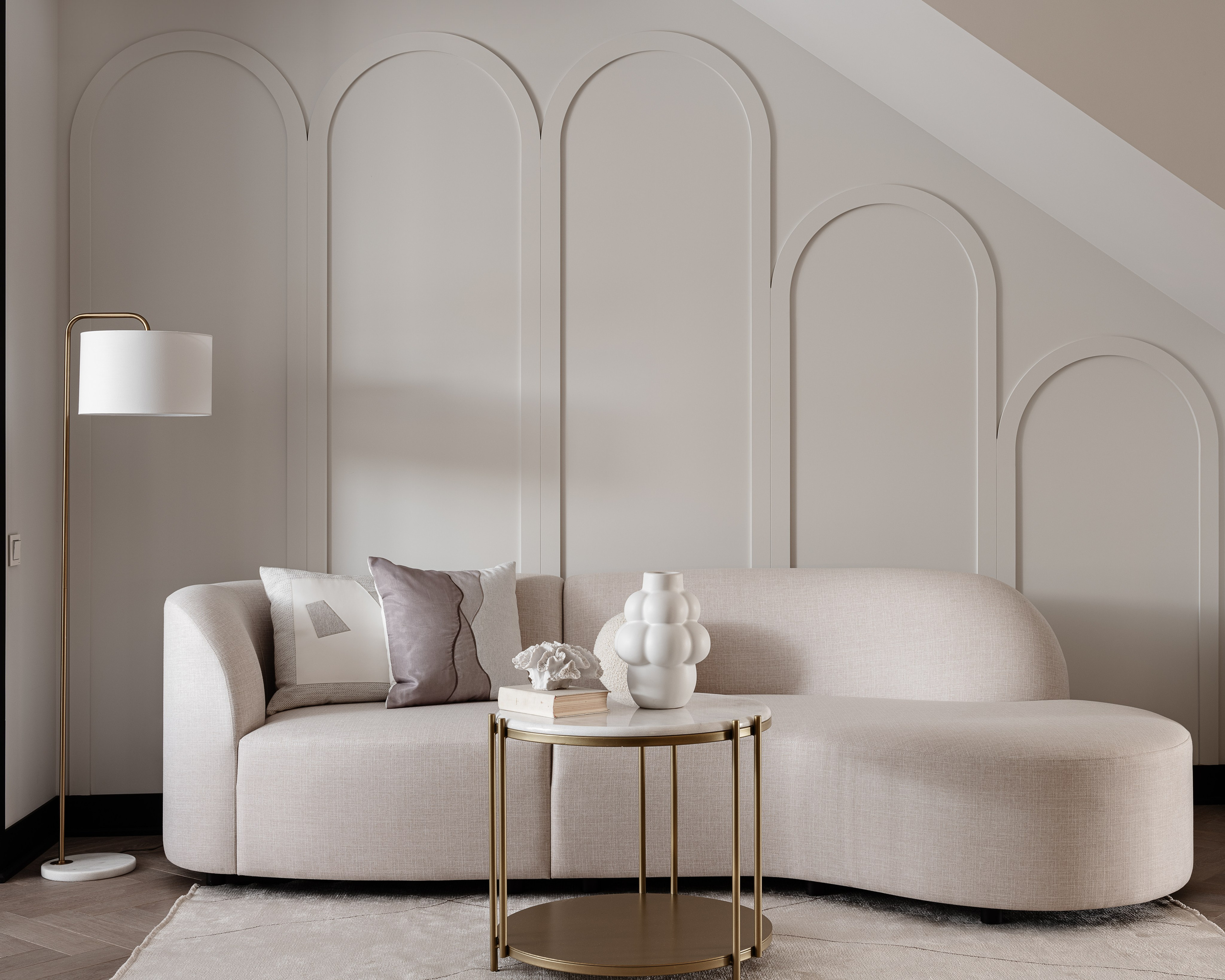 Современный интерьер гостиной с изогнутым диваном в молочном оттенке, журнальным столиком на золотом каркасе и декором в пастельных тонах. Стена украшена арочными панелями, автор Василисса Бачева, стилист Наталья Якушева, фото Сергей Денисенко.