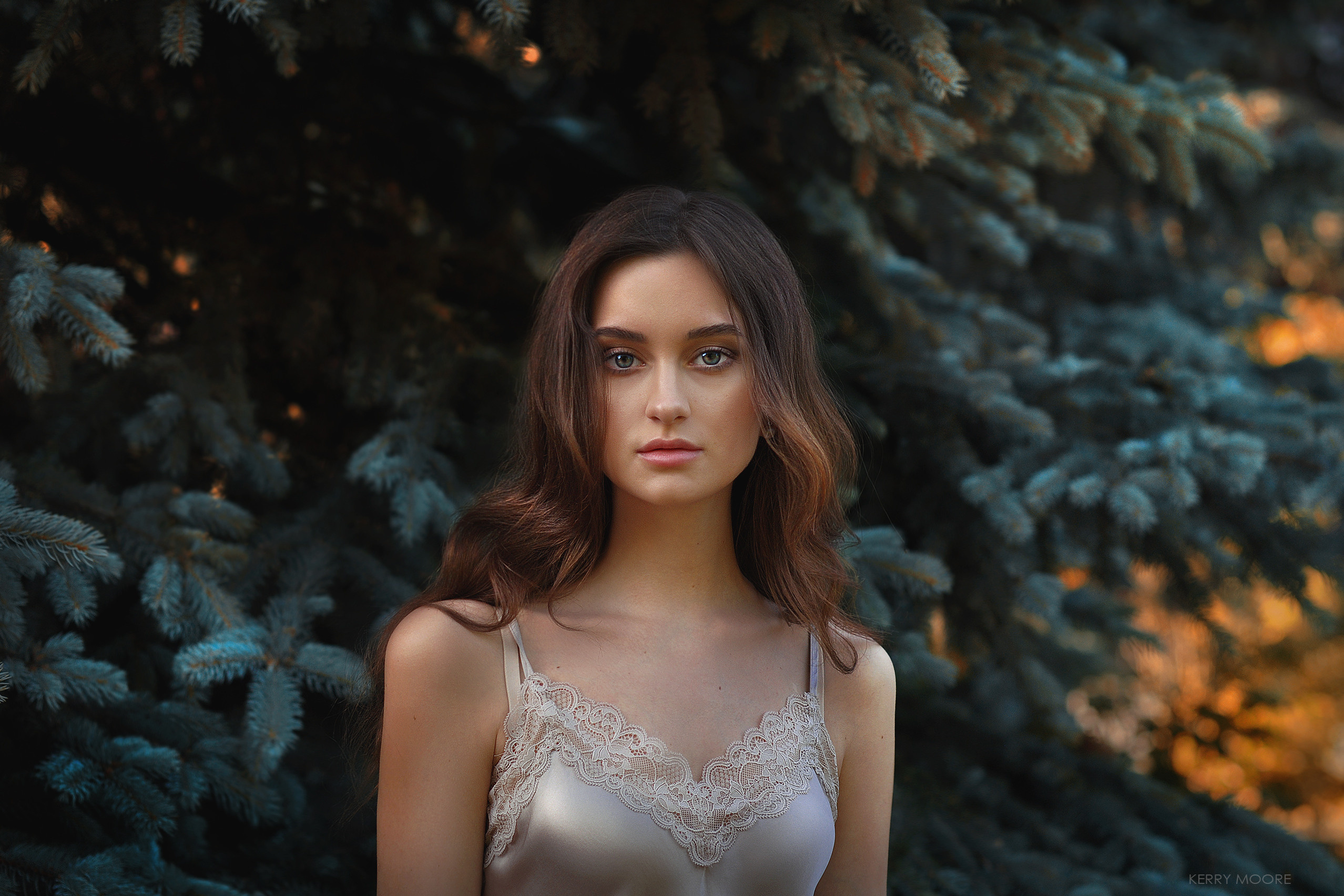 Красивая девушка стоит в шёлковой кружевной майке возле голубой ели