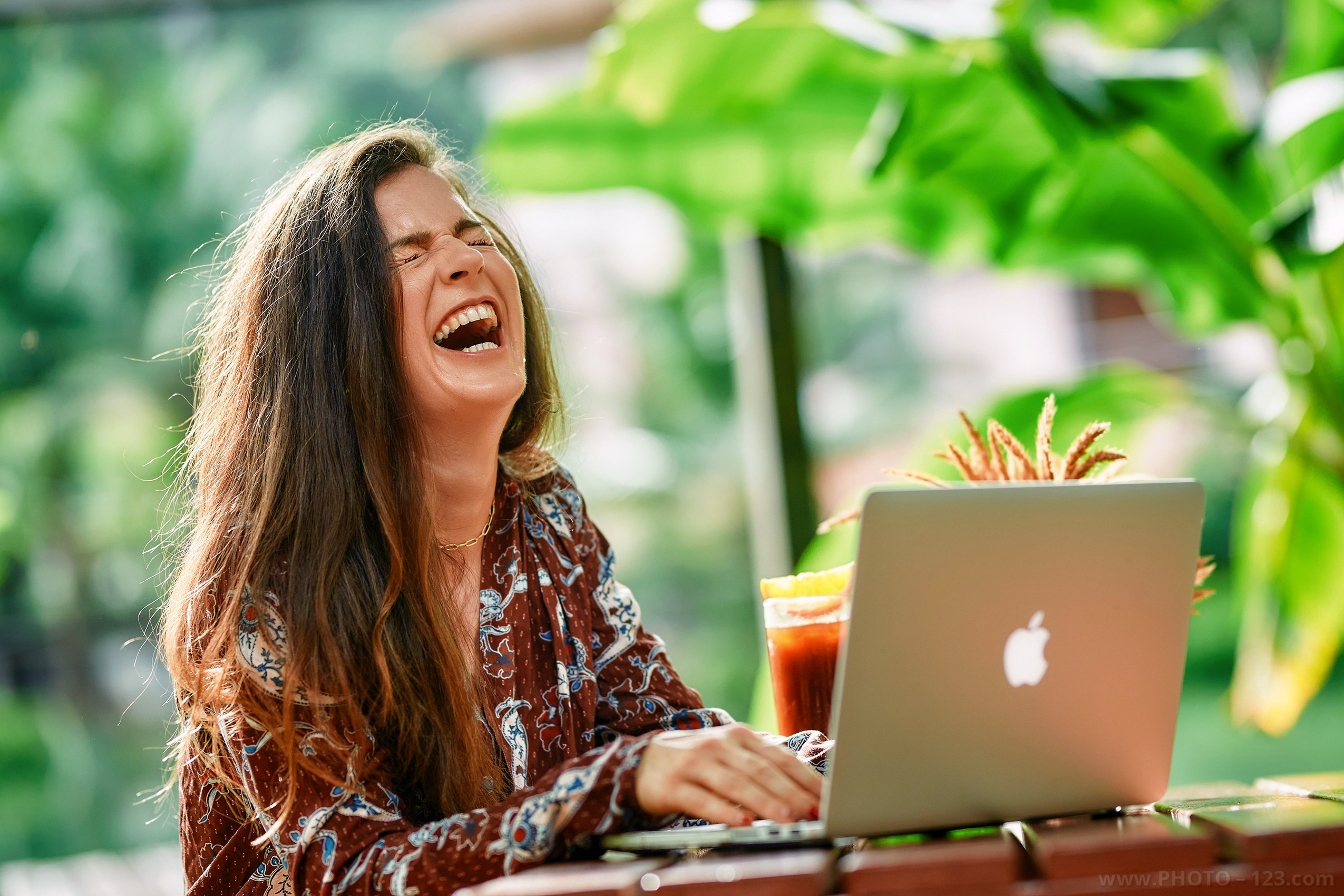 Lifestyle portrait of joyful young woman laughing while working on laptop in tropical garden cafe, remote work lifestyle photography in Phu Quoc, natural light portrait, relaxed atmosphere, professional photographer Vietnam