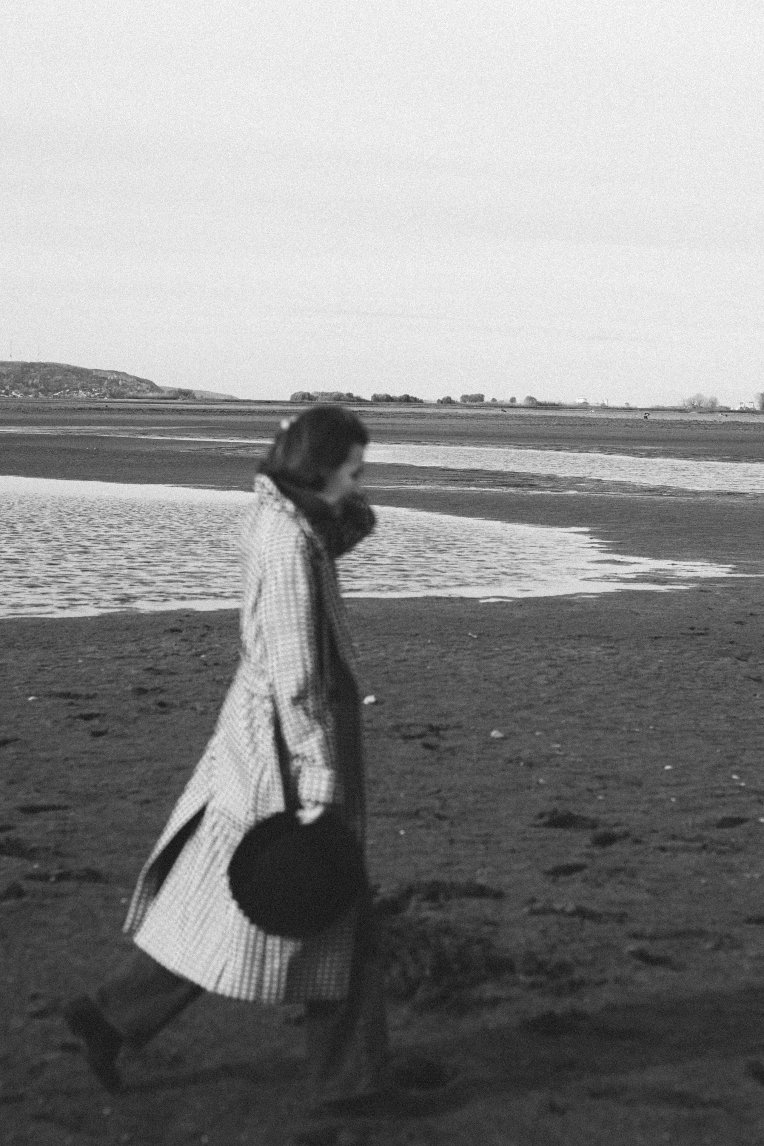 Женщина в клетчатом пальто идет по пляжу, держа шляпу.