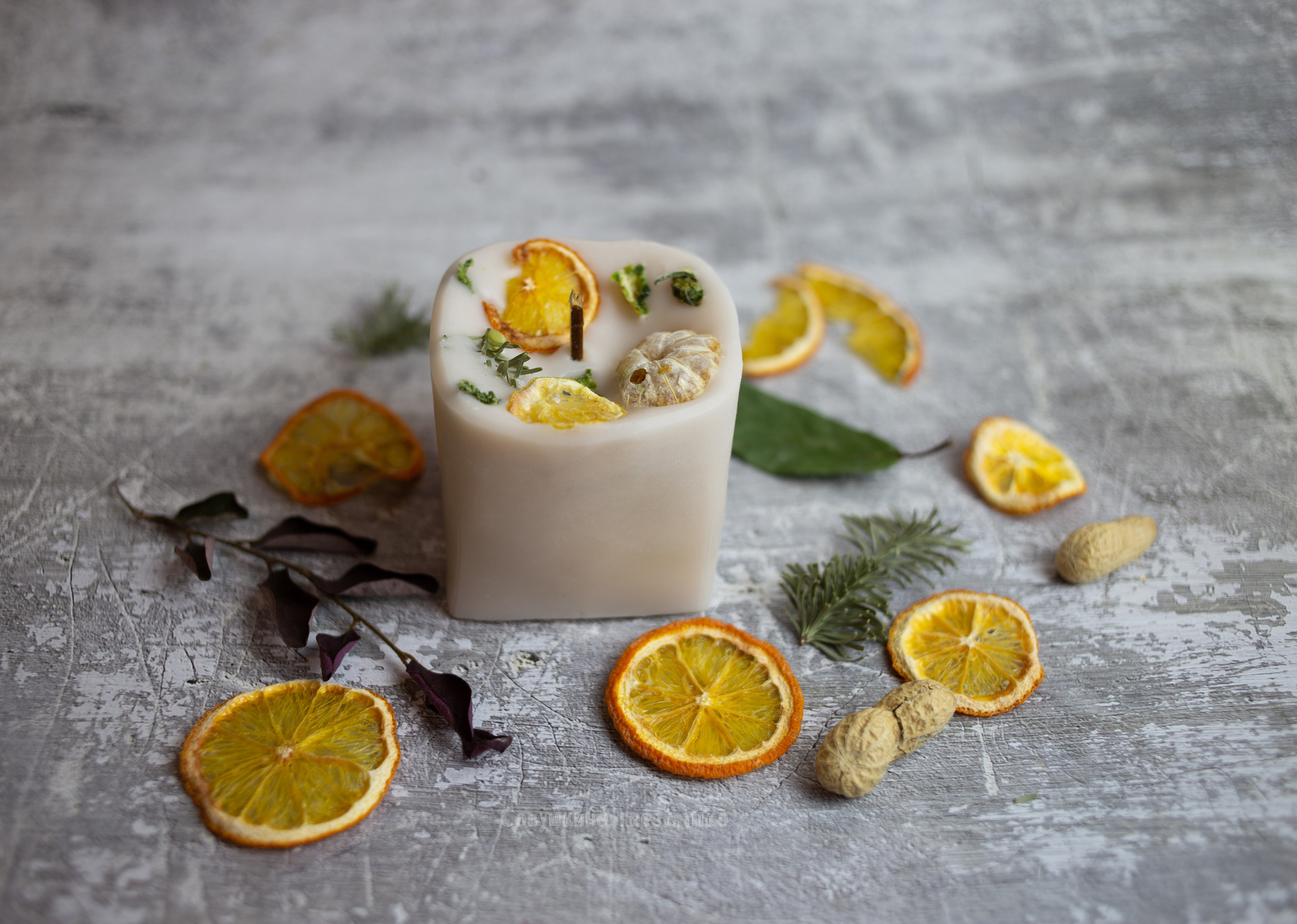 квадратная   свеча ручной  работы, украшенная  апельсинами. на  сером фоне. рядом  лежат сушенные  апельсины.