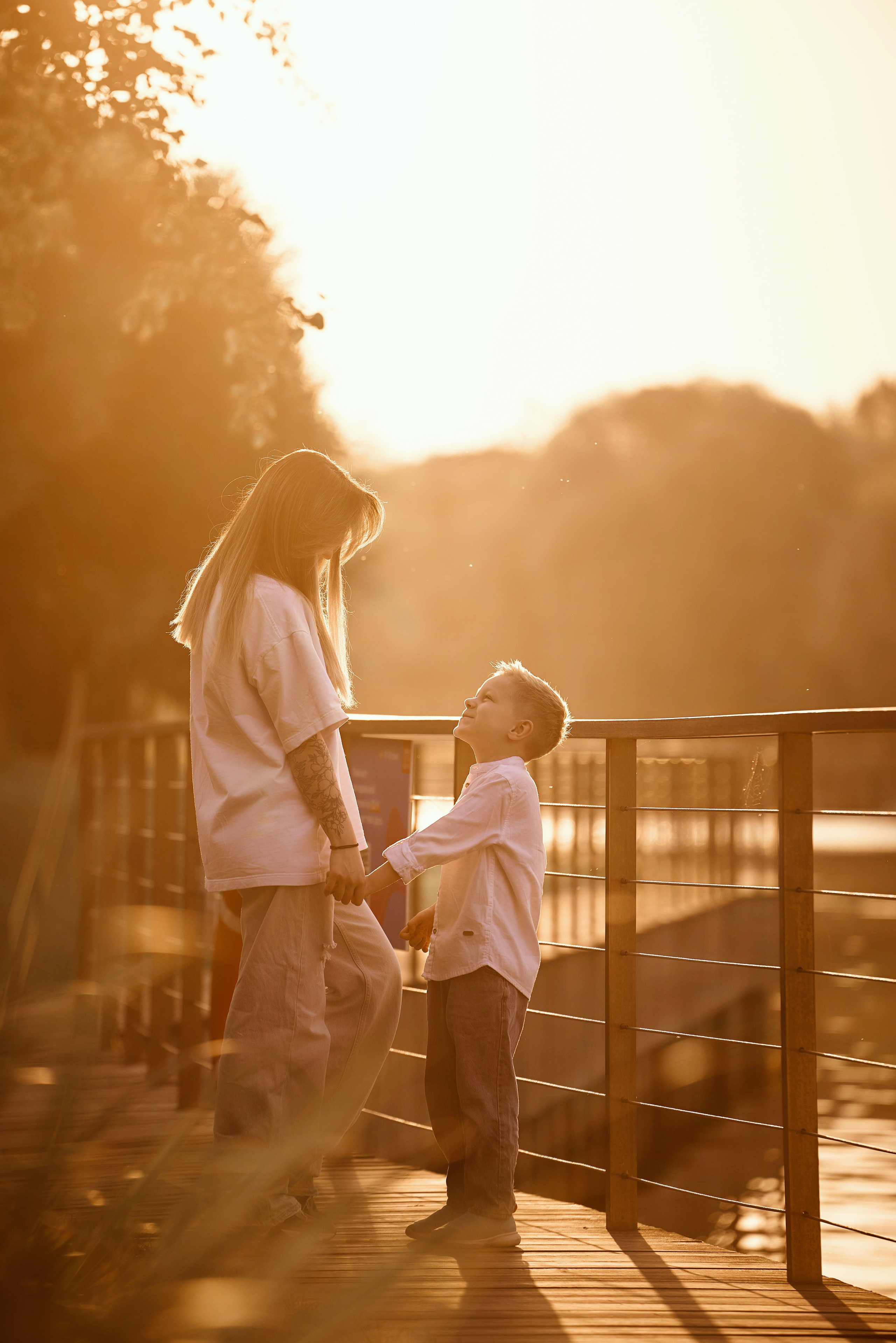мама с мальчиком в закатном солнце у пруда на ВДНХ держуться за руки и смотрят друг другу в глаза, девушка в белой рубашке и голубых джинсах, мальчик в белой рубашке и голубых джинсах