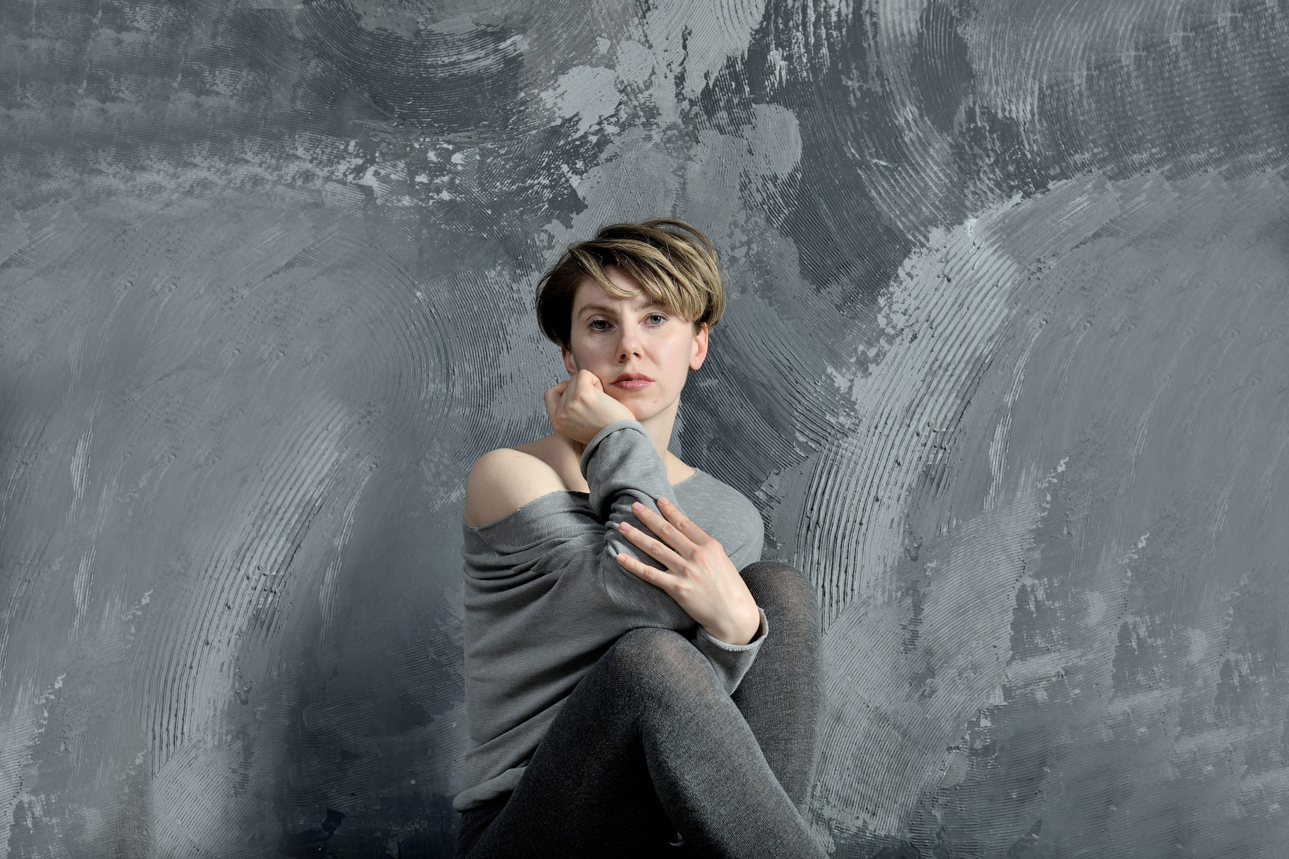 Девушка в сером свитере сидит на фоне бетонной стены в фотостудии.