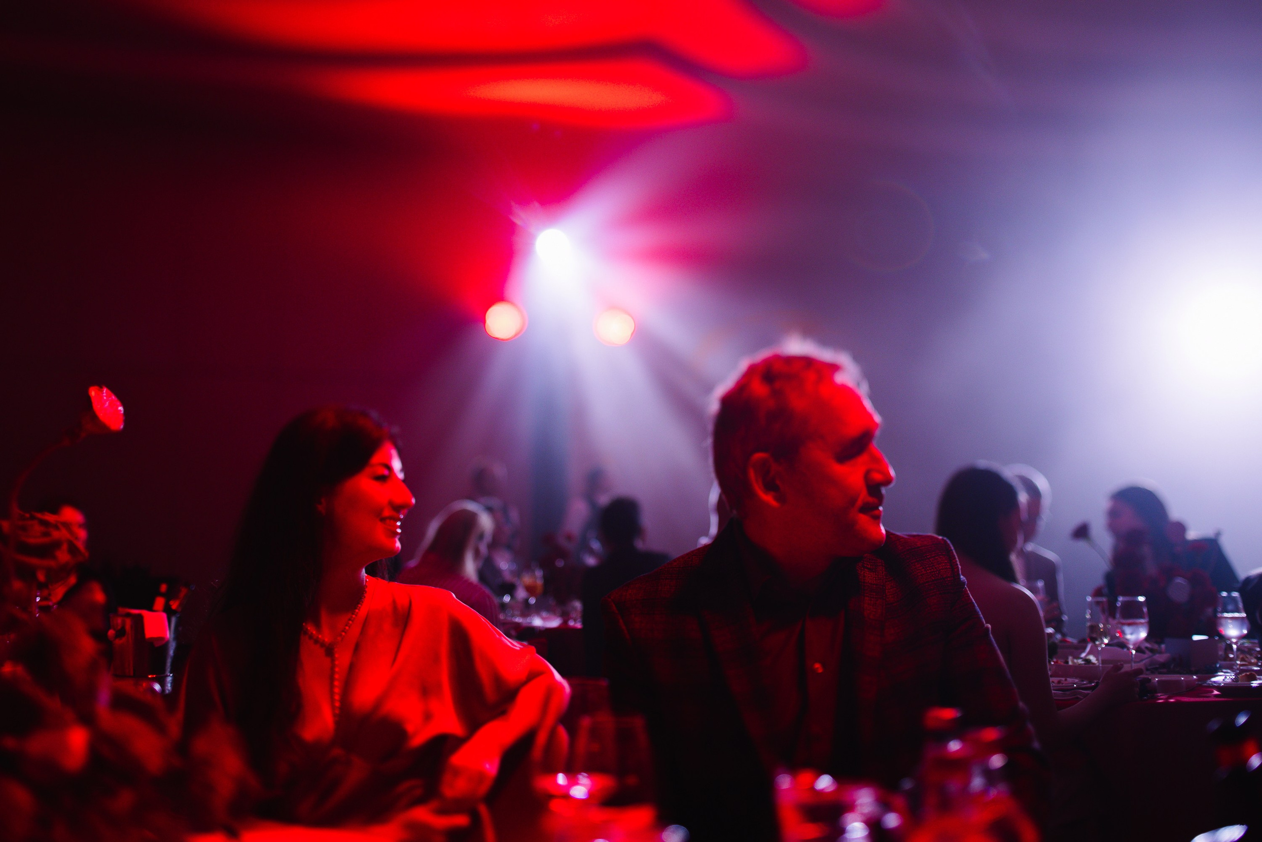 Гости вечера: Мужчина и женщина за столиком в зале, освещенном розовыми и синими лучами прожекторов.