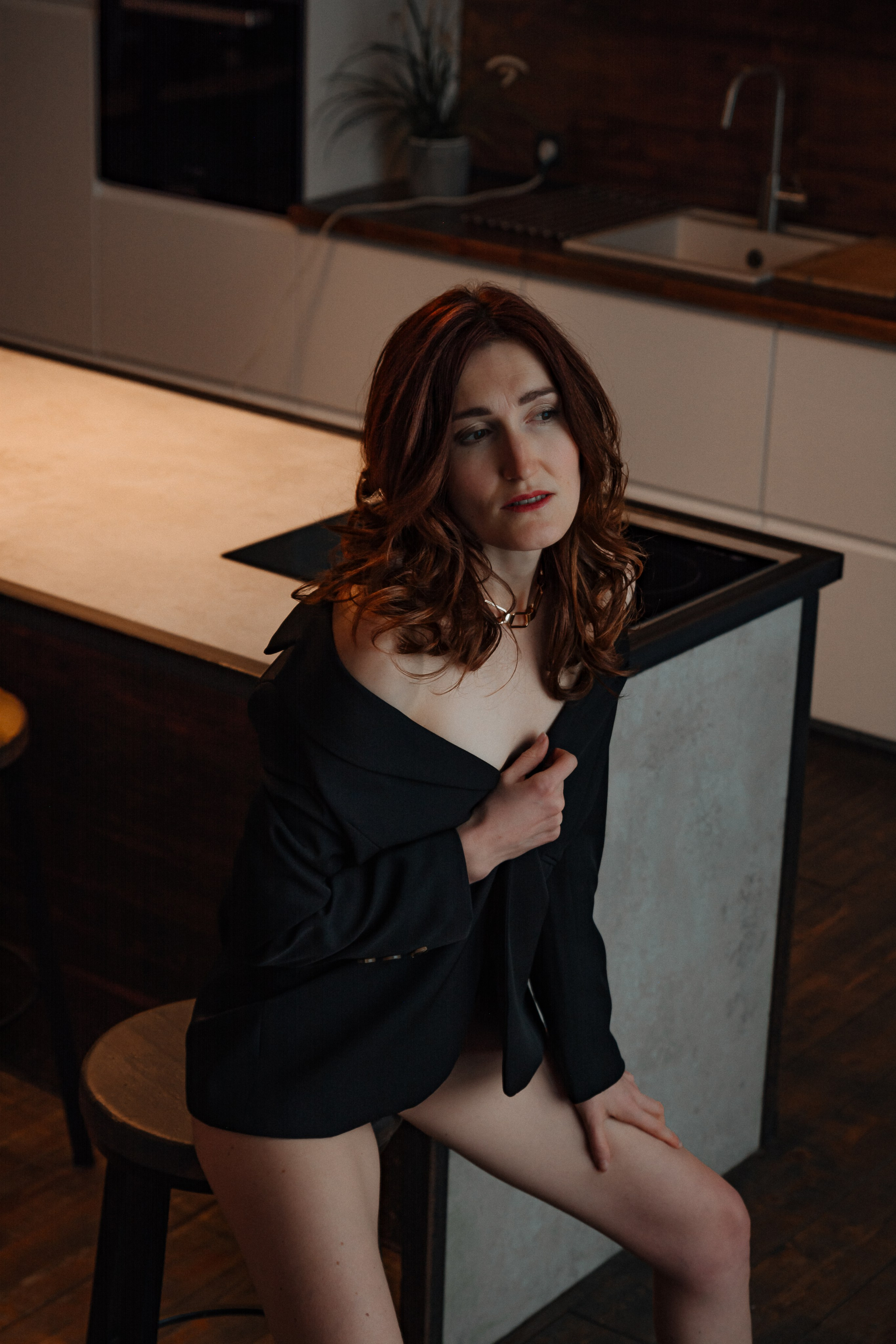 Девушка придерживает полы пиджака, слегка приоткрывая соблазнительный вид на гладкие стройные ноги. В полумраке комнаты ее кожа сияет бархатным блеском.