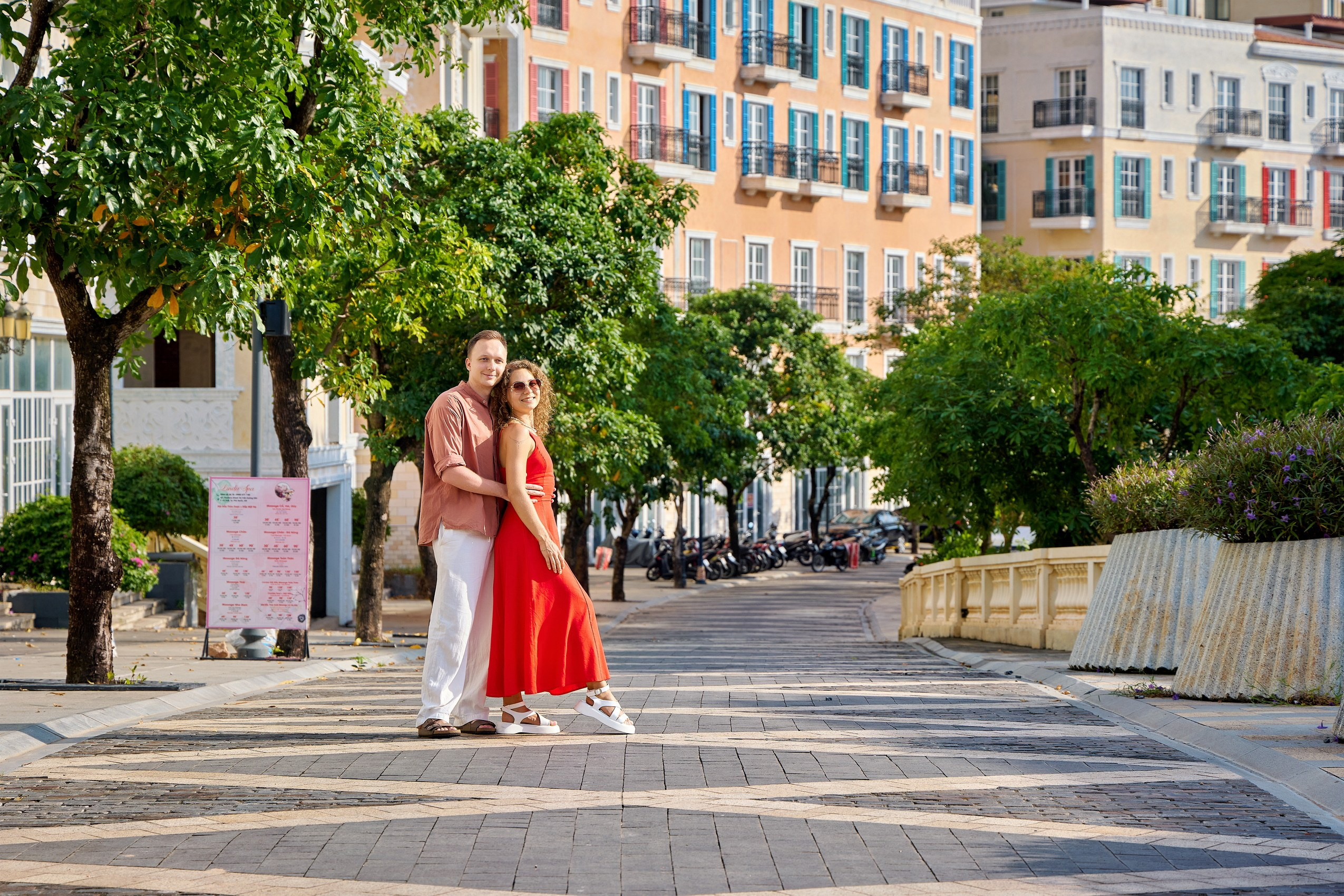 Sunset Town couple photoshoot on tree‑lined street with pastel buildings and parked motorcycles, Phu Quoc, Vietnam.