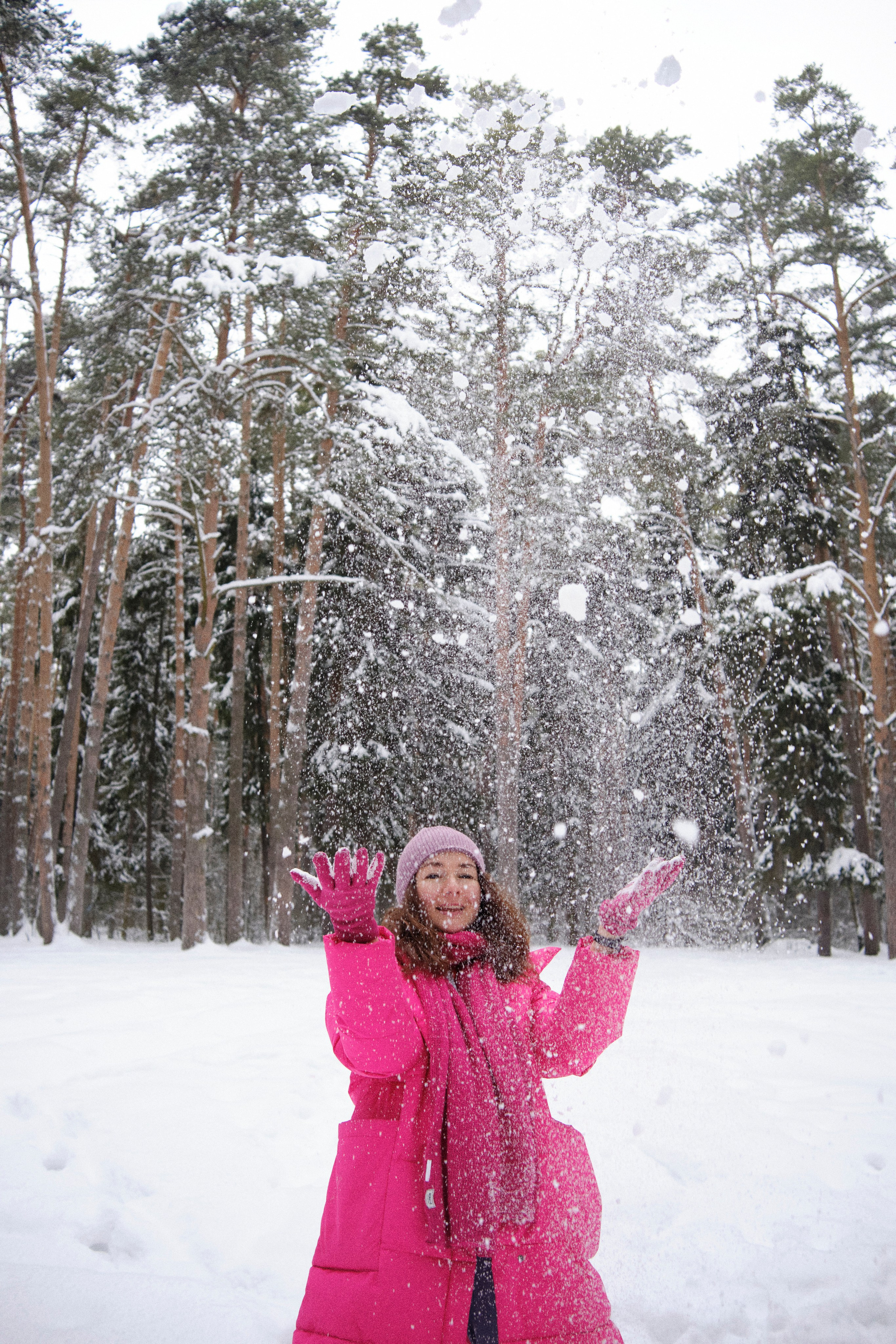 Девушка в яркой одежде подбрасывает снег в зимнем сосновом лесу