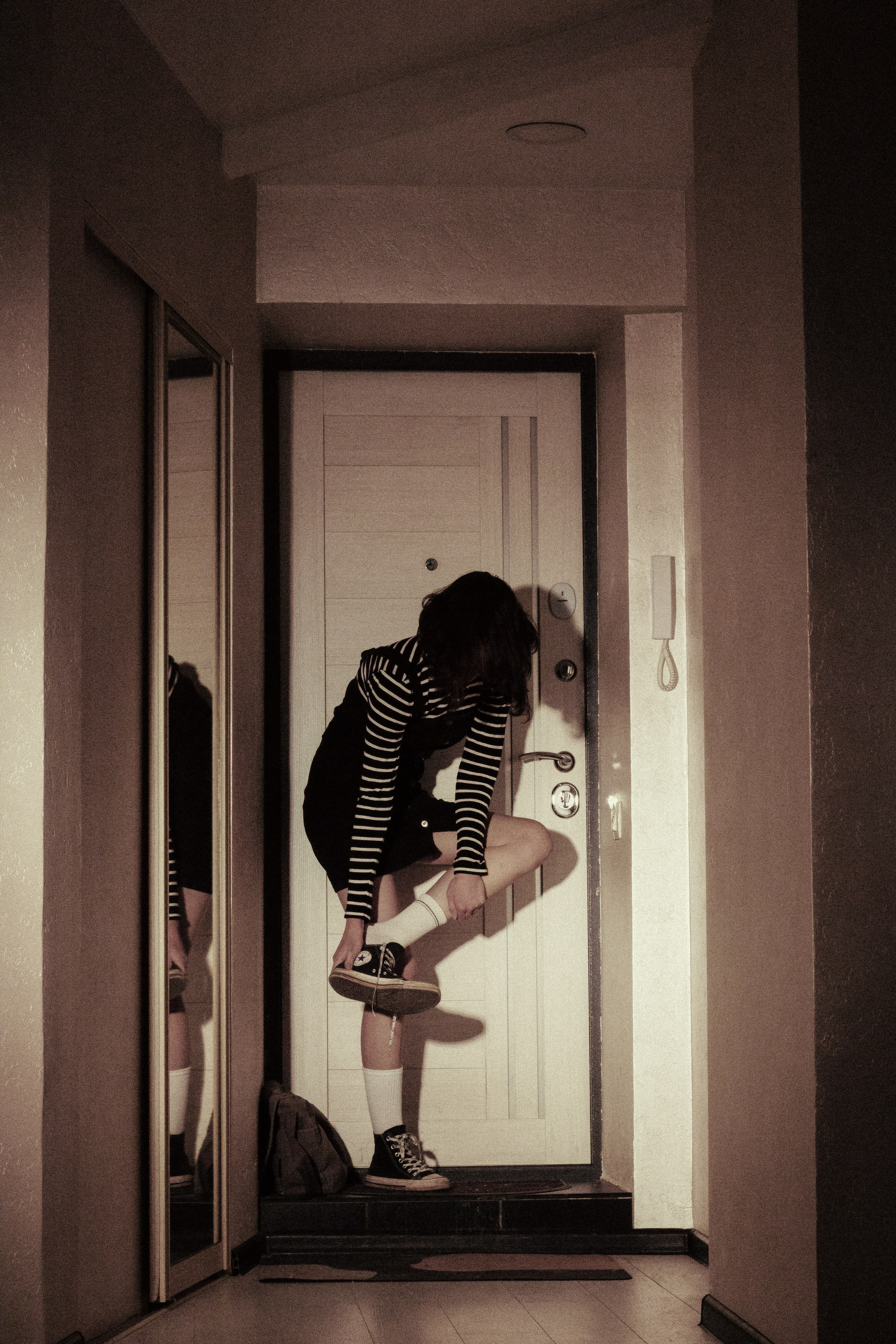 Девушка в конверсах наклоняется у входной двери, завязывая шнурки — готовясь войти туда, где её уже не ждут.