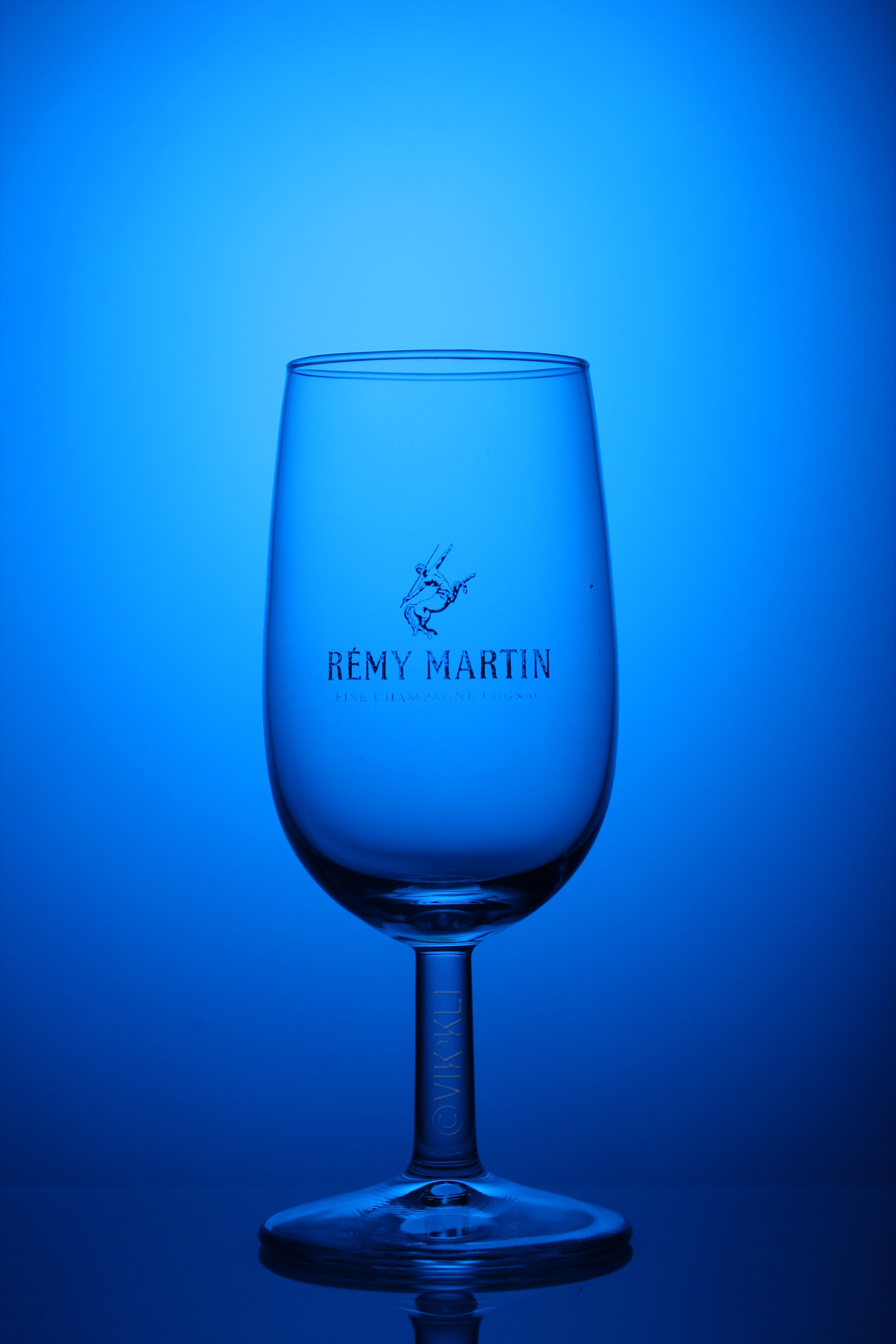 Стеклянный бокал Remy Martin на синем фоне. Предметное фото.
