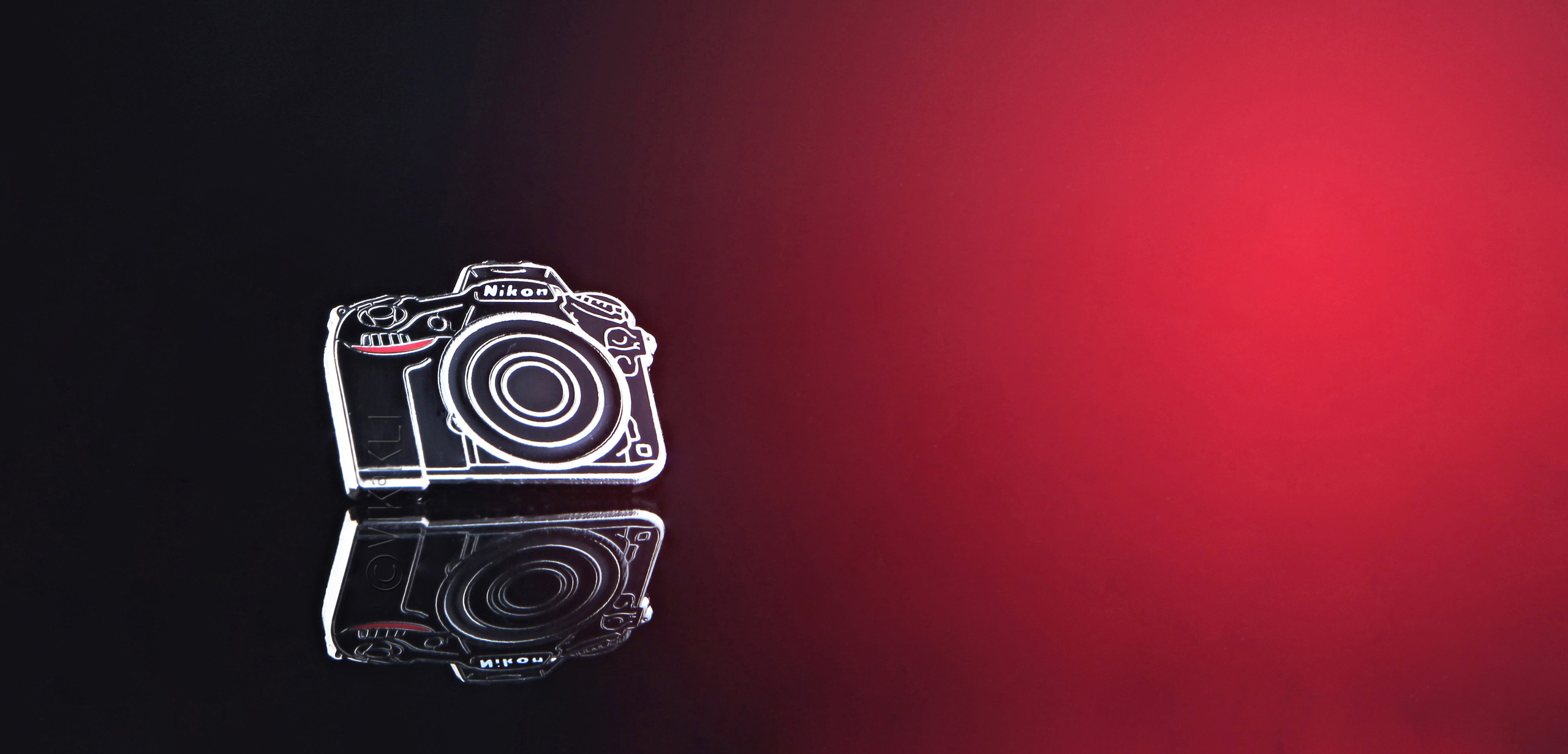 Металлический значок фотоаппарат Nikon на тёмном фоне. Предметное фото.