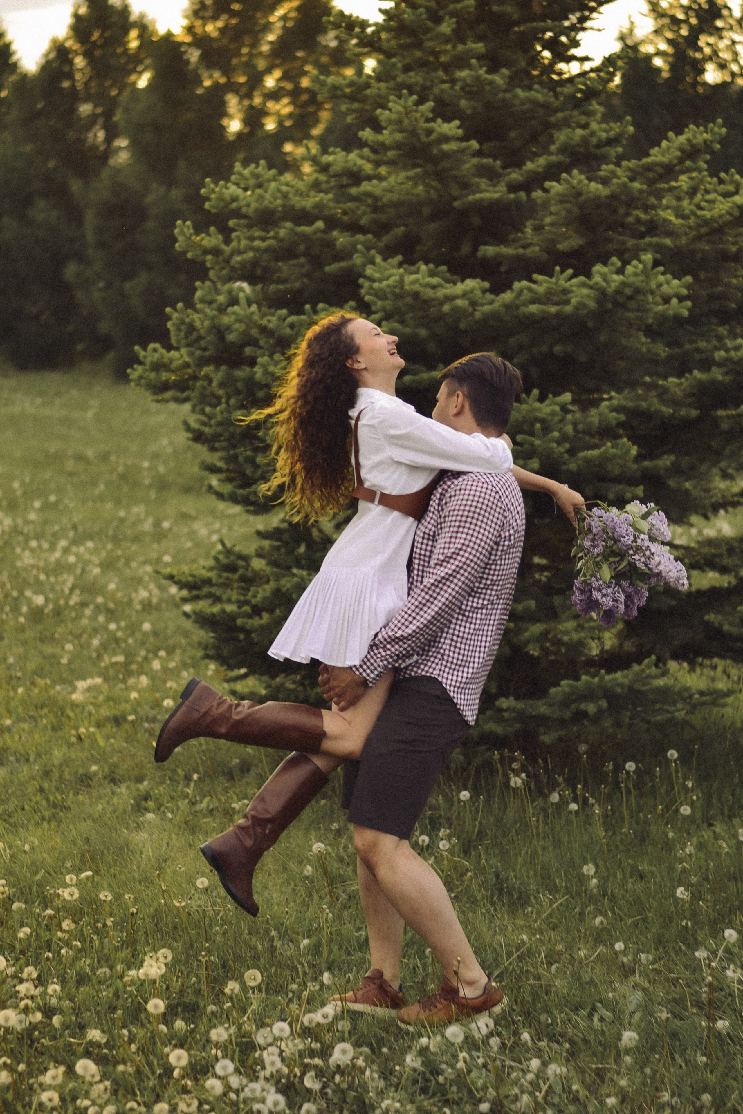На фотографии изображена пара, которая, кажется, наслаждается моментом счастья и любви на природе. Они стоят среди зелёной травы, окружённые деревьями и кустарниками, что создаёт атмосферу уединения и покоя. Женщина в белой блузе и джинсовой юбке с поясом, обутая в высокие коричневые сапоги, держит в руках букет фиолетовых цветов, вероятно, лаванды. Мужчина в клетчатой рубашке и шортах обнимает её, и они оба улыбаются и смотрят вверх, словно наслаждаясь солнечным светом или чем-то приятным. Их позиция и выражения лиц говорят о том, что они испытывают радость и беззаботность.