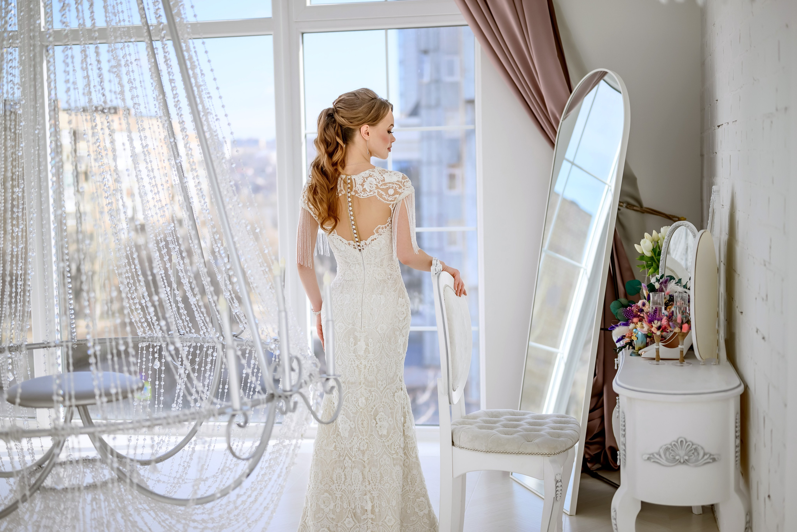 Невеста в белом платье с красивыми пуговицами на спине стоит возле большого зеркала