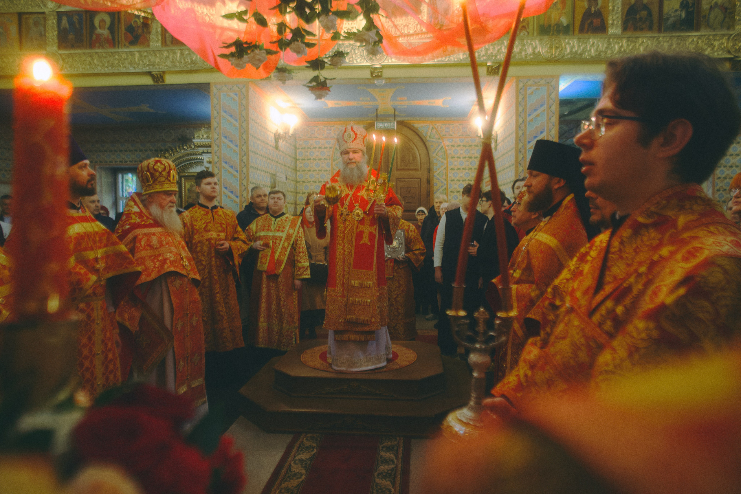 Архиерей в митре и красных облачениях стоит на возвышении посреди храма во время пасхального богослужения. Вокруг — духовенство и прихожане.