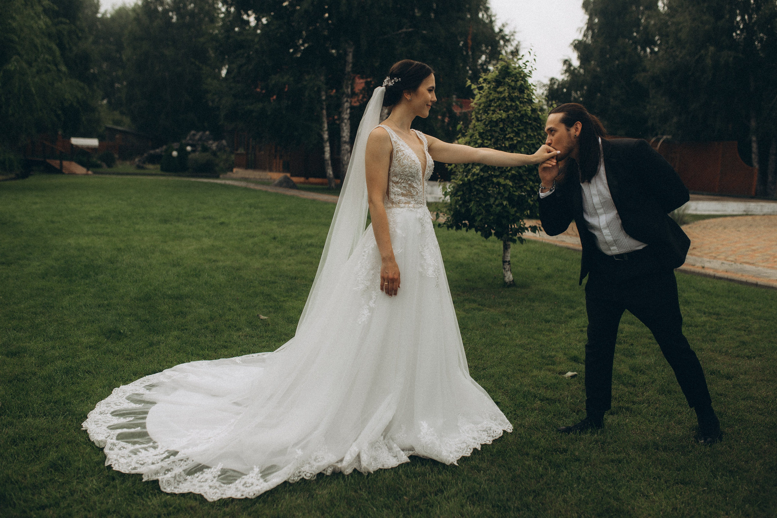 Романтический жест: жених Никита целует руку невесты Линды на зеленом газоне. Свадебная классика.