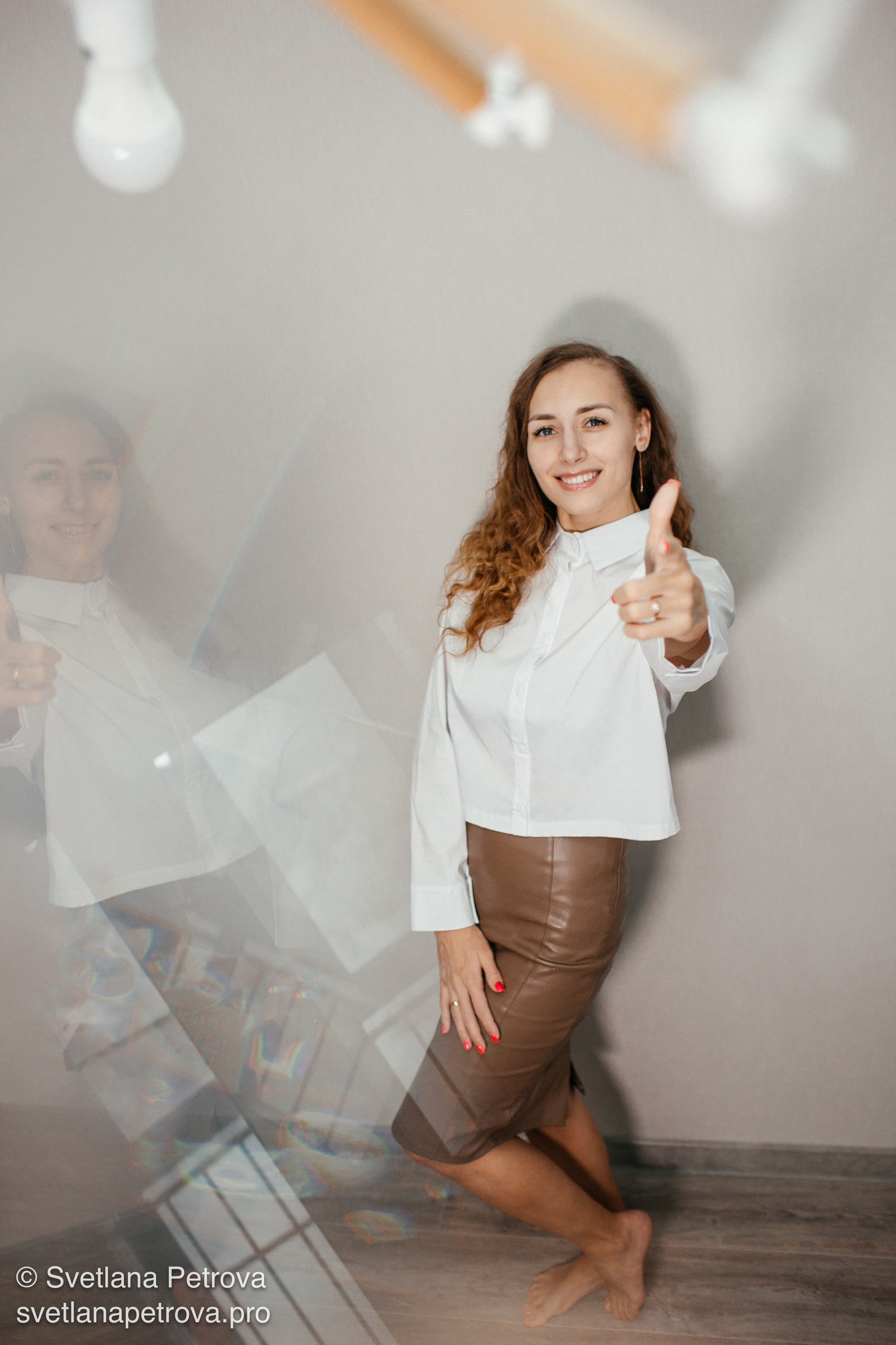 Улыбающаяся девушка в белой блузке — красивый портрет. Модный образ: белая рубашка, кожаная юбка. Девушка с «палец вверх» — энергичный кадр. Портрет в студии: стиль, улыбка, поза с жестом. Элегантная поза, светлая блузка, коричневый юбка-карандаш. Студия, девушка босиком, позитивный настрой. Стильный образ — улыбка и жест одобрения.