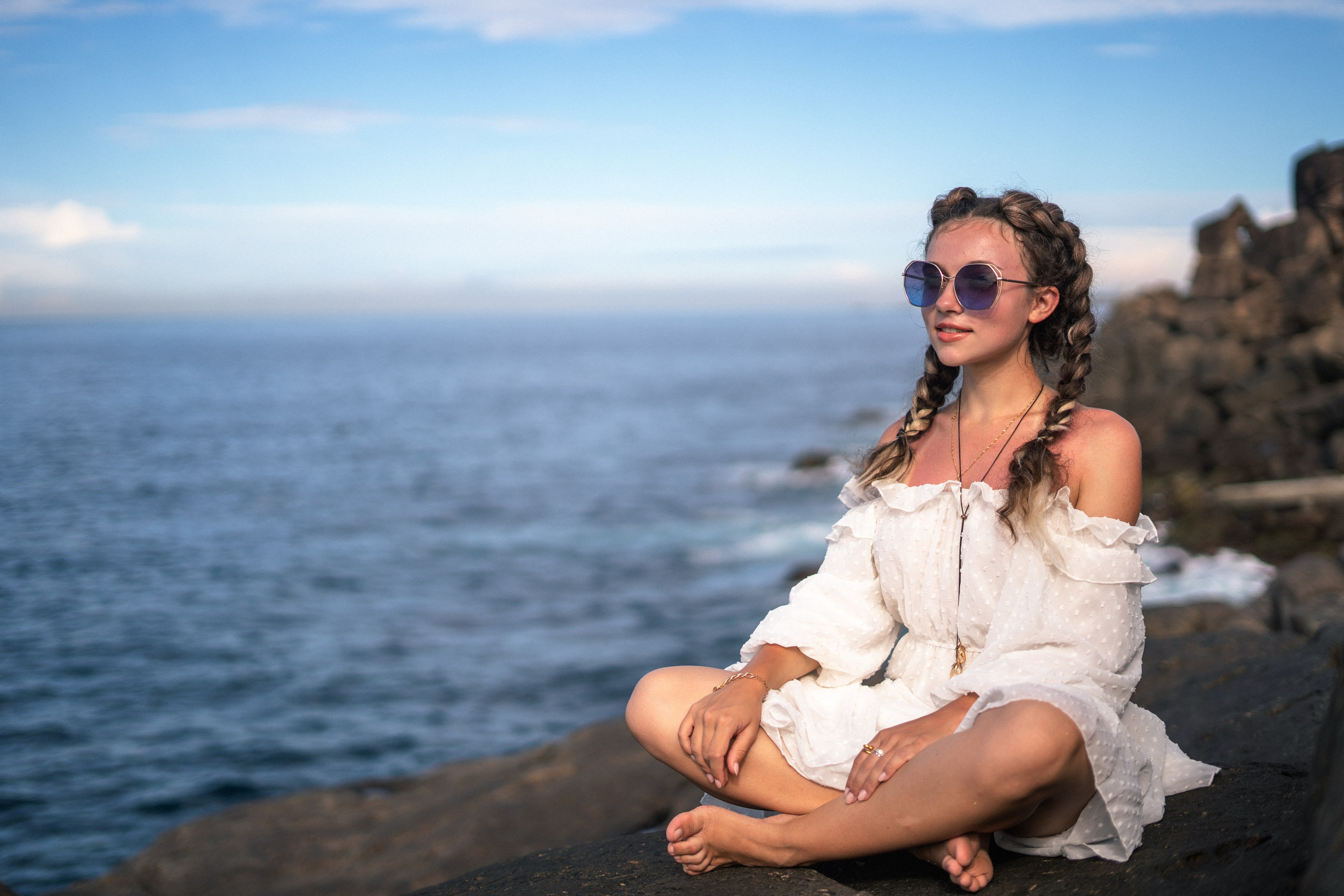 a girl in a white dress and glasses with the sunset by the ocean in the background