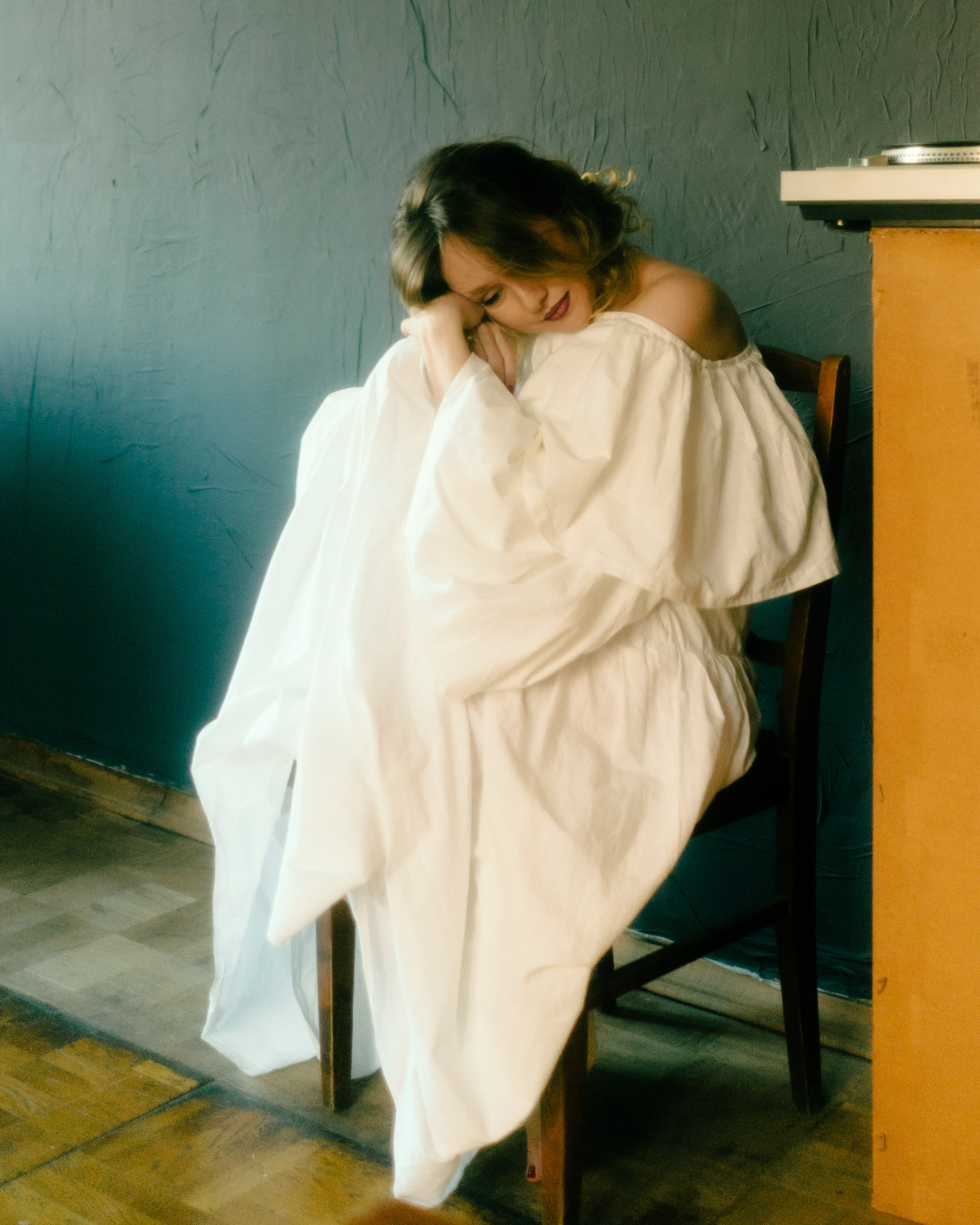 Девушка в объемном белом платье на стуле у серой стены — чувственная фотосессия беременности в ретро-фотостудии Санкт-Петербурга.