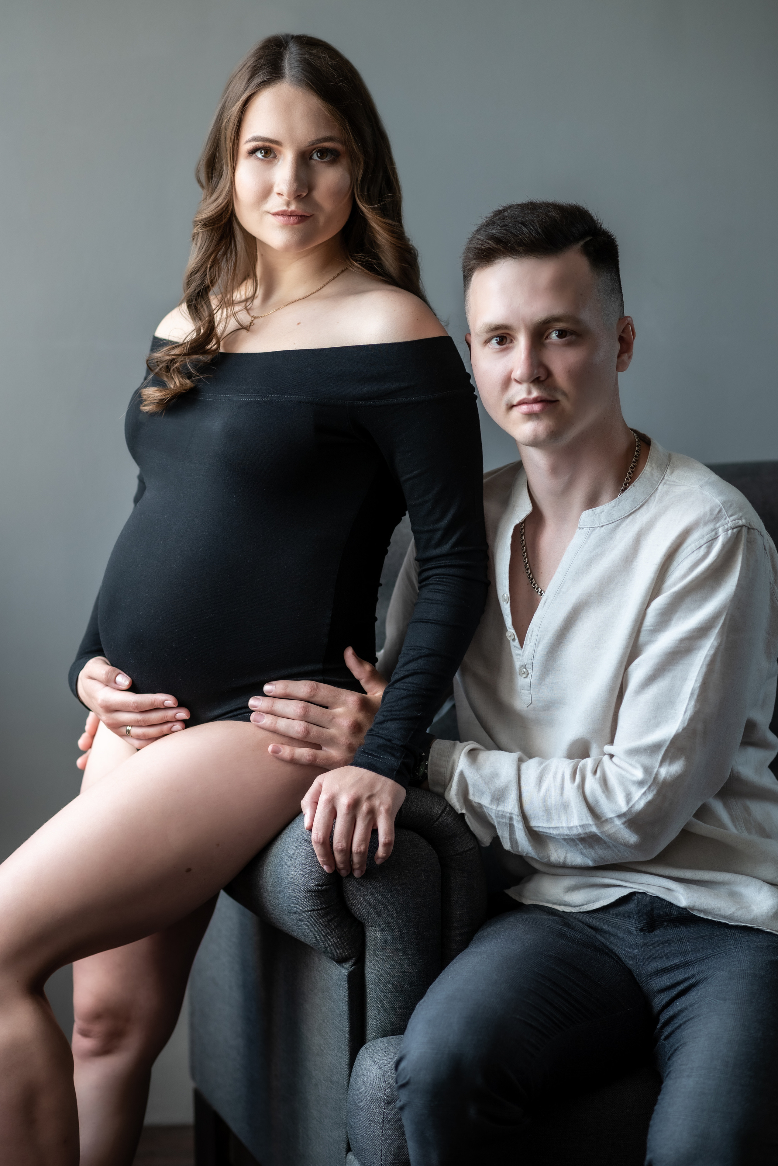 беременная женщина сидит на подлокотнике серого дивана рядом с мужем