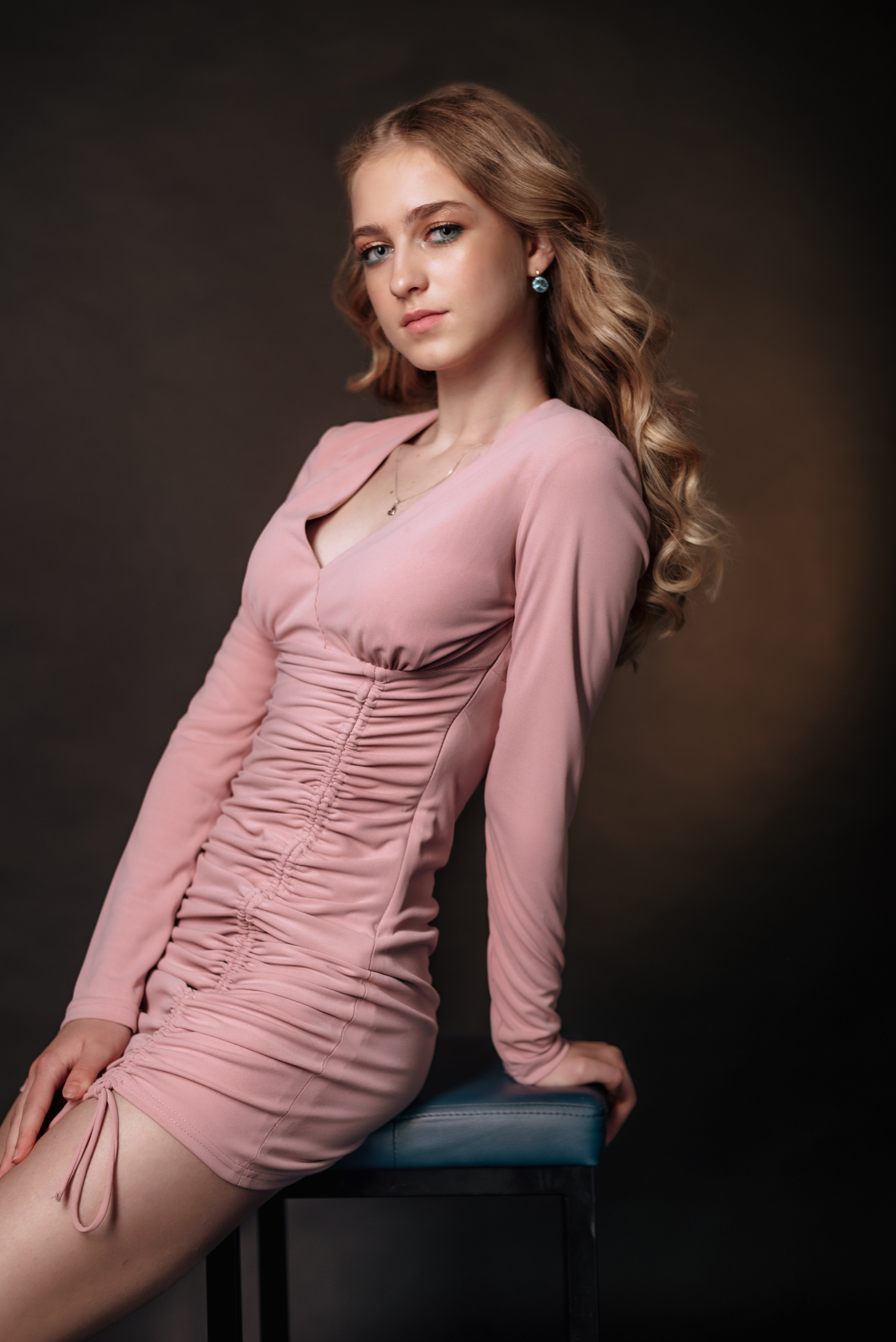 юная девушка в пыльно-розовом платье на черном фоне сидит на высоком табурете