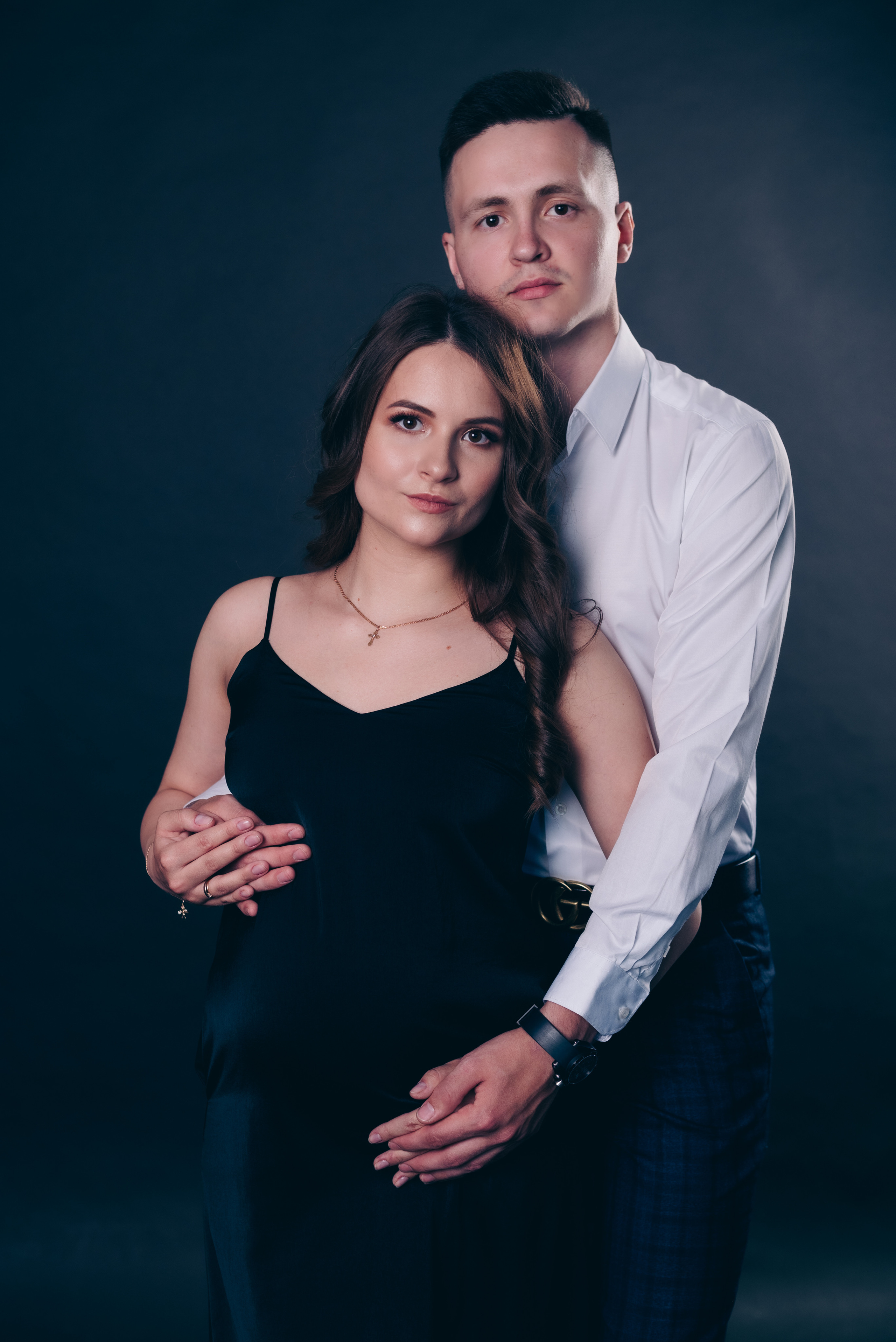 муж обнимает беременную жену в атласном черном платье сзади, поясной портрет