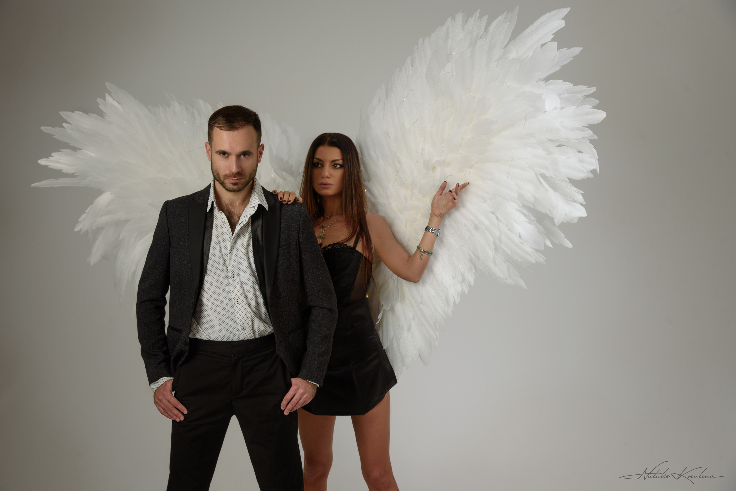 мужчина в деловом костюме стоит с руками в кармане женщина с крыльями ангела в фэшн стиле держит его за плечо