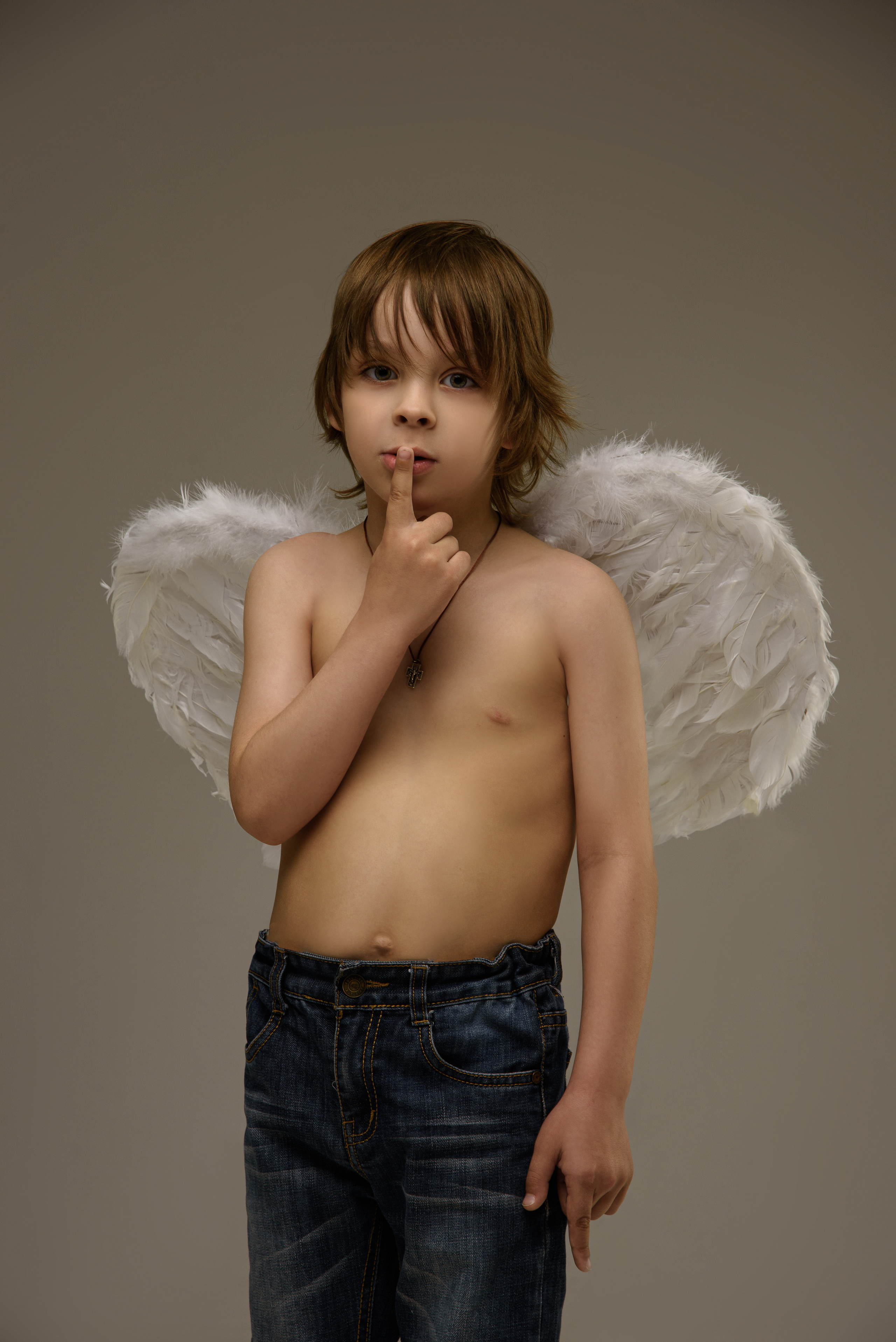 мальчик ангел в джинсах с крыльями за спиной прижимает палец к губам