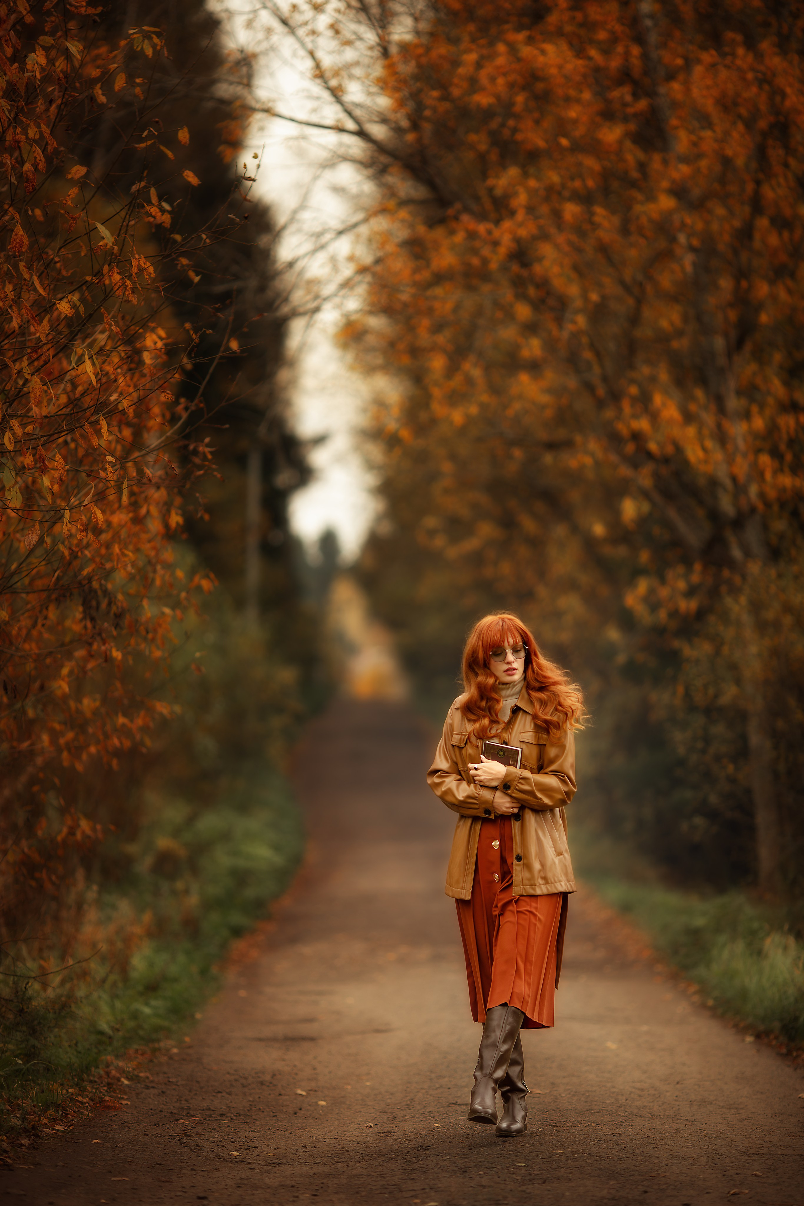 Девушка с рыжими волосами в осеннем пальто идёт по аллее с золотыми листьями.