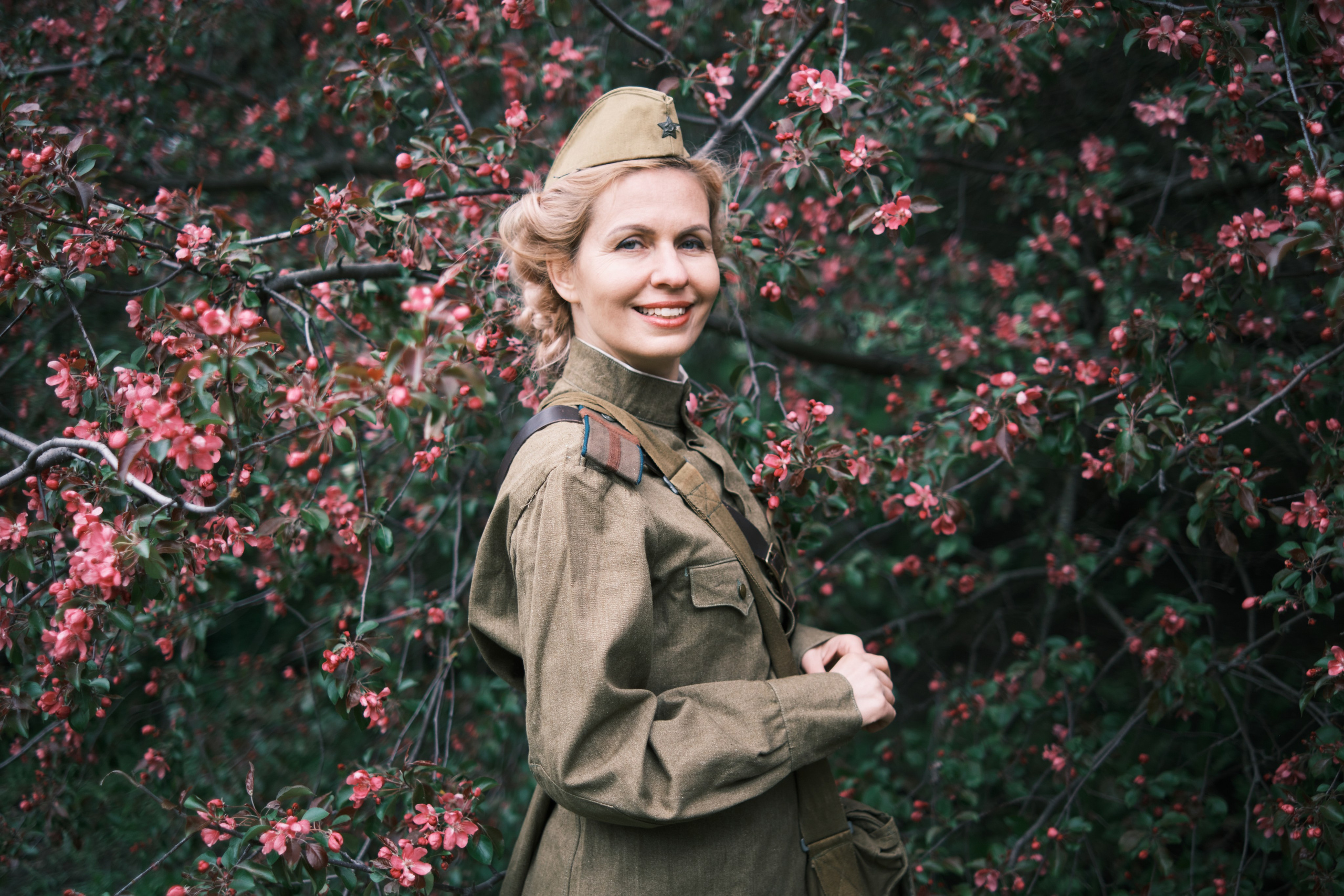 Девушка в военной форме и пилотке стоит на фоне цветущих яблонь