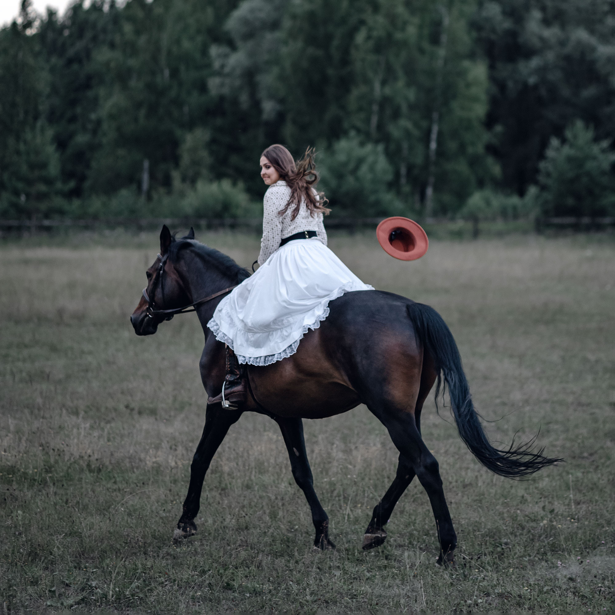 Девушка в длинной юбке скачет по летнему полю на лошади карей масти. Волосы развеваются на ветру, шляпа улетает.