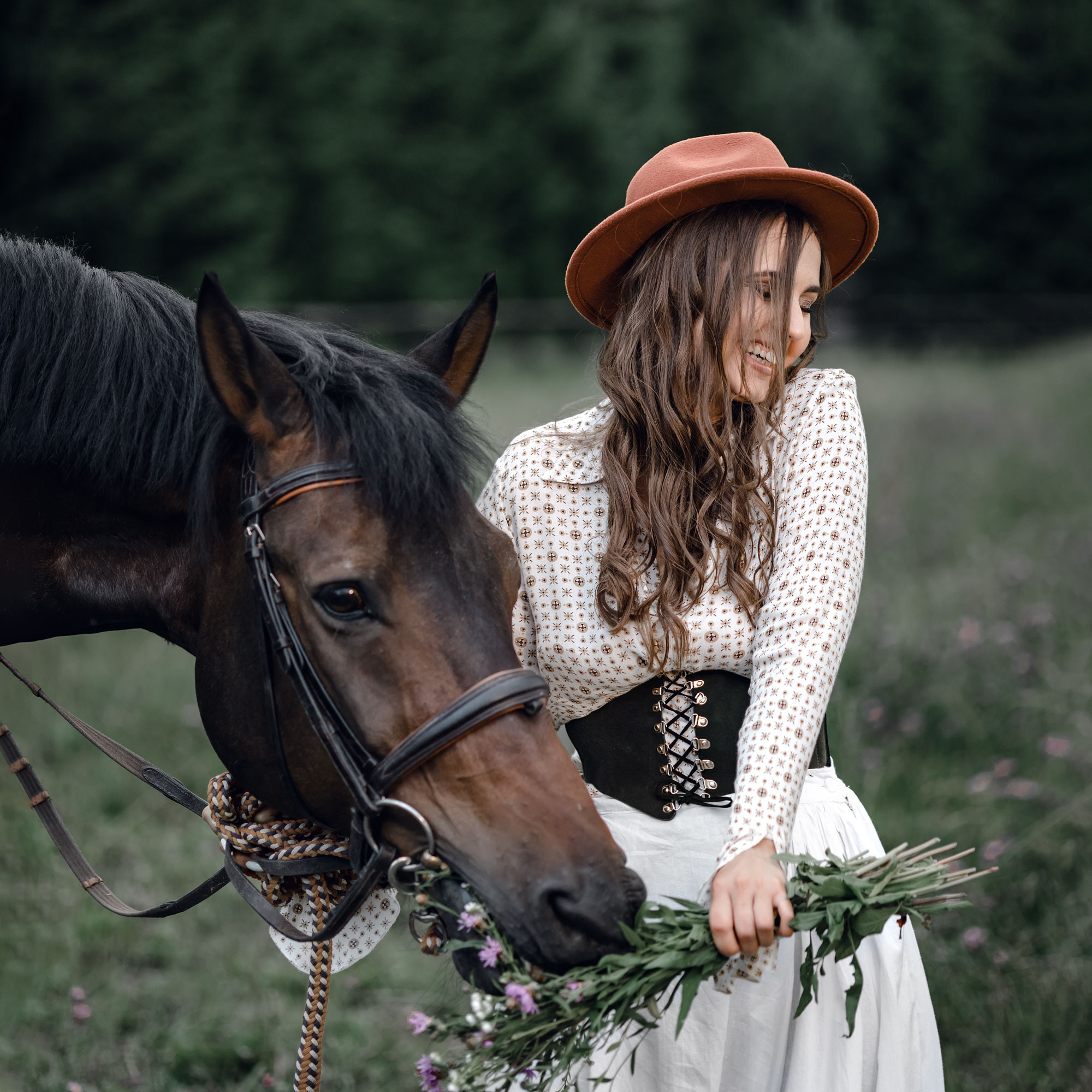 Лошадь ест букет цветов, который держит в руках девушка в шляпе. Летняя фотосьемка с лошадью, девушка смеется.